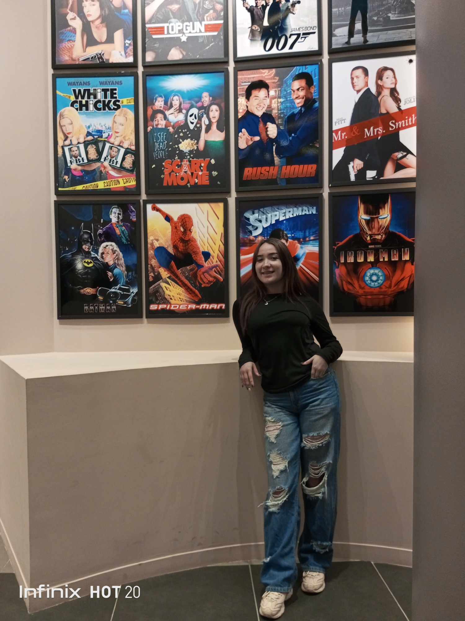 A young woman stands smiling in an indoor setting, posing against a light-colored wall adorned with framed movie posters. She has long, dark brown hair, parted in the middle, and is wearing a dark green long-sleeved top, ripped light blue jeans, and white sneakers. Her right hand is in her pocket, and her left arm rests on a white ledge that runs along the bottom of the wall.
The wall behind her features a grid of nine movie posters arranged in three rows of three. From top left to bottom right, the visible posters include:
* Pulp Fiction (partially visible, top left)
* TOP GUN
* James Bond 007 (likely Die Another Day or similar)
* Mr. & Mrs. Smith (partially visible, top right)
* WHITE CHICKS
* SCARY MOVIE
* RUSH HOUR
* BATMAN (featuring Batman and Joker, likely the 1989 film)
* SPIDER-MAN (classic comic book style)
* SUPERMAN (classic comic book style, behind the woman)
* IRON MAN (behind the woman)
The woman is positioned slightly to the right of the frame, facing the camera with a gentle smile. The lighting suggests an indoor, well-lit environment, possibly a cinema lobby, a game room, or an entertainment venue. The bottom left corner of the image contains a watermark: "Infinix HOT 20". The floor is dark with visible tile lines. The overall impression is a casual, relaxed pose in a fun, movie-themed environment in Porlamar, Venezuela.