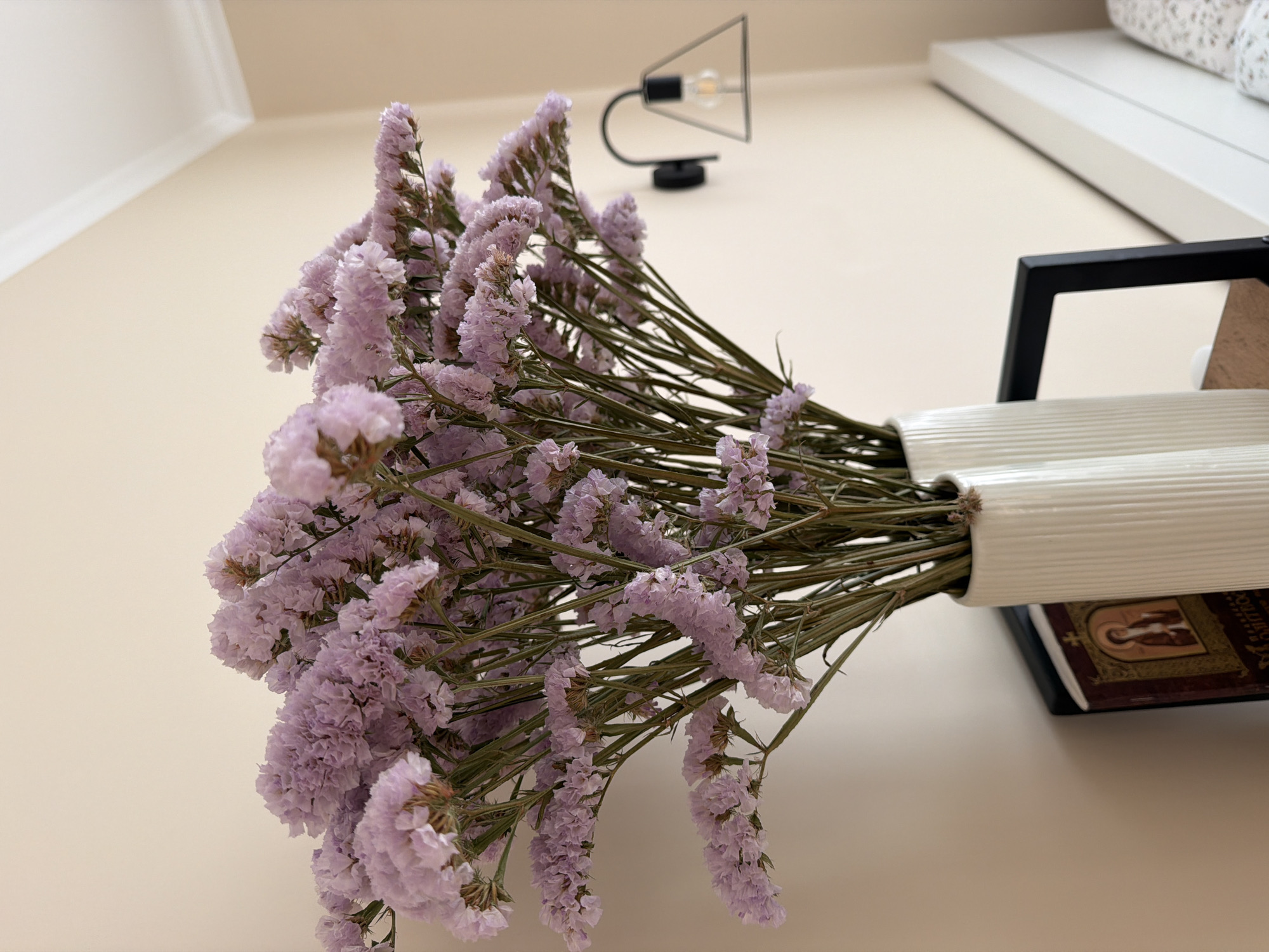 A bouquet of dried lavender statice flowers is displayed in a fluted white vase on a surface, possibly a nightstand. The flowers are a light purple color and appear delicate. Behind the vase, to the left, is a book with a religious image on its cover. To the right of the vase, a black framed object is partially visible. Further in the background, a wall-mounted light fixture with a geometric metal shade and an exposed bulb is present. The wall is a neutral, light beige color, and the ceiling has white molding. A portion of a white headboard and patterned bedding are visible on the far right of the image, suggesting the scene is in a bedroom. The lighting in the image is soft and natural, likely from ambient room light. There are no people visible in the image.