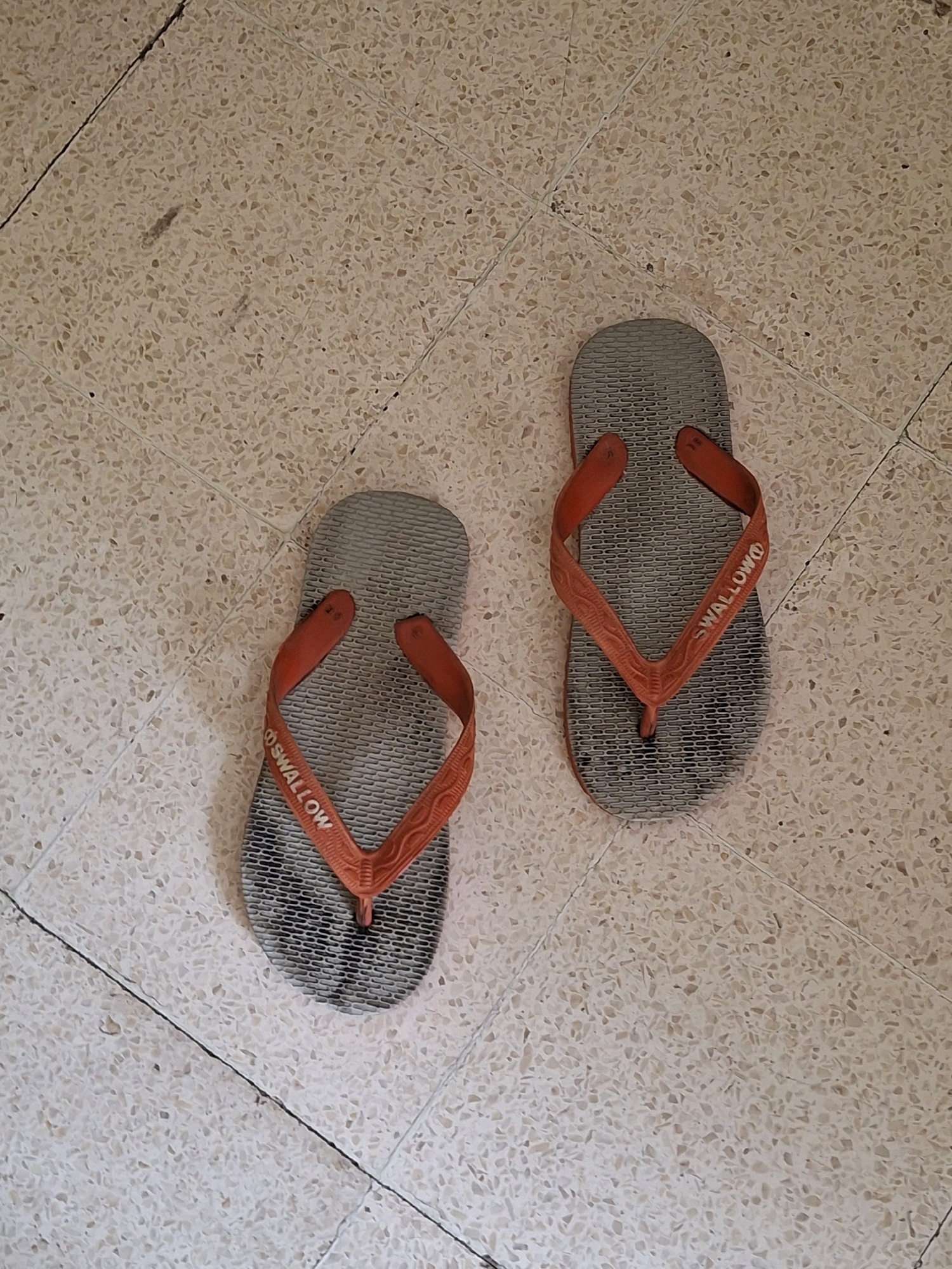 Two grey flip-flops with orange straps are lying on a tiled floor. The flip-flops are positioned next to each other. The flip-flop on the left is angled with the toe pointing towards the lower left corner of the image, and the flip-flop on the right is positioned with the toe pointing towards the bottom center of the image. The tiled floor appears to be in a house or building in Lumajang, Indonesia. The tiles are beige and speckled with small darker particles. There are grout lines between the tiles, forming a grid pattern.