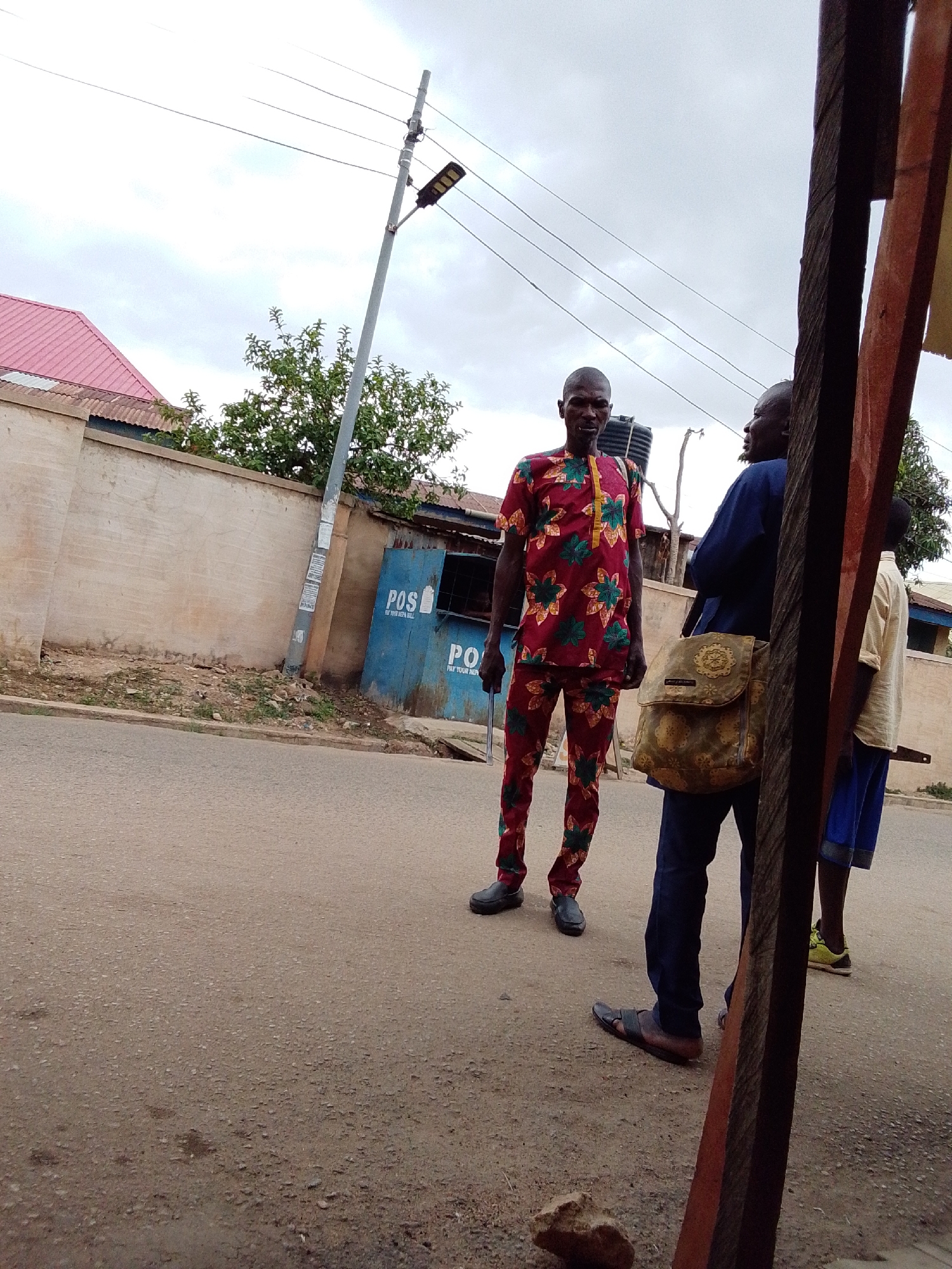 The image captures an outdoor street scene featuring three men. In the foreground, a man stands centrally, facing the camera directly. He is dressed in a distinctive red and green patterned traditional shirt and matching trousers, and wears dark shoes. A small black tank-like object is visible behind his head, seemingly worn on his back.
To the right of the central figure, partially obscured by a dark wooden frame in the extreme foreground, another man is visible from the back. He wears a dark blue long-sleeved top and trousers, and carries a prominent gold and green patterned messenger-style shoulder bag. He is wearing dark sandals. Further to the right, a third person is partially seen, wearing a light-colored top, blue shorts, and bright yellow-green sneakers.
The setting is a paved road with a curb. To the left, a light-colored concrete wall stretches along the roadside, topped by a building with a reddish corrugated iron roof. A tall utility pole with a modern LED street light and several overhead power lines dominates the background on the left. Further down the street, two blue kiosks are visible, clearly marked with the text "POS" and "PAY YOUR NEPA BILL." Sparse green foliage and trees are seen behind the wall and kiosks. The sky is overcast, indicating a cloudy day.