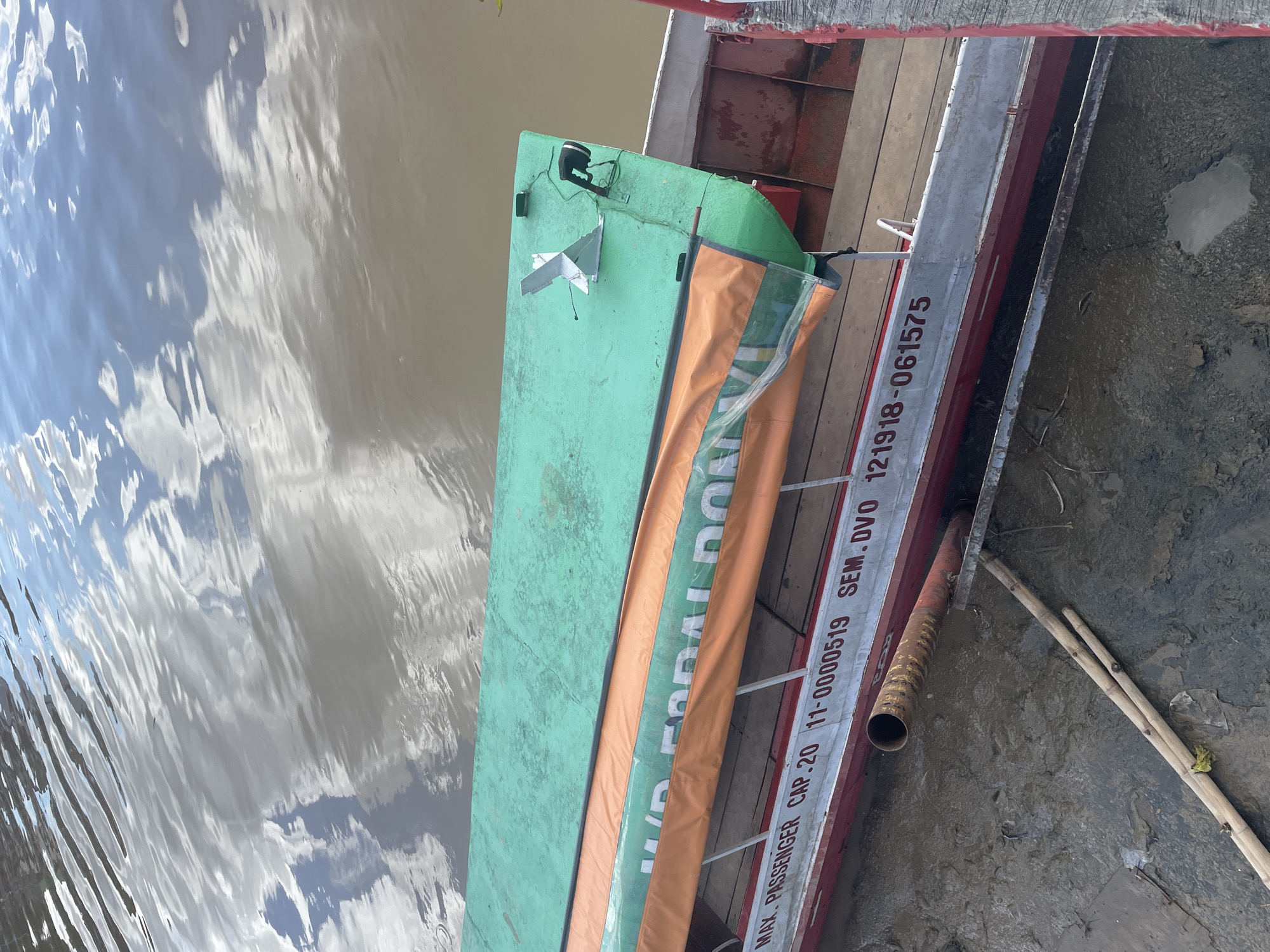 The image captures a section of a boat docked on a muddy riverbank, likely in Davao City, Philippines, given the "SEM. DVO" marking. The boat, painted red and white with a long, green roof, has an orange cover draped over its side, bearing white lettering that is partially obscured but includes "MAX. PASSENGER CAP. 20". A metal anchor or similar fixture is visible on the green roof, along with a black light and wiring. The water in the background reflects the sky and clouds, appearing somewhat murky. On the riverbank, several bamboo poles rest against the boat, and the ground is composed of dark, wet mud with some debris. The perspective is from above, looking down at the boat and its immediate surroundings.