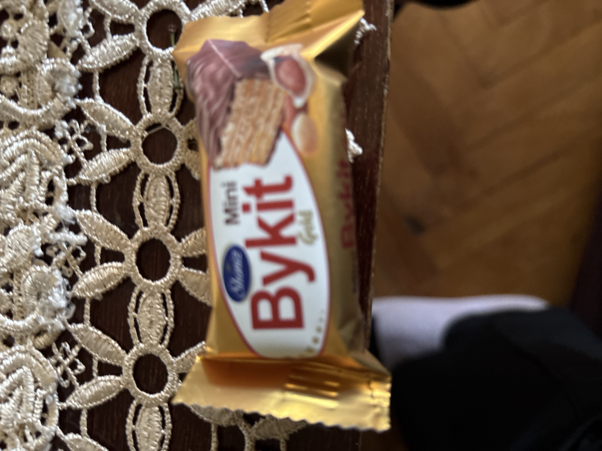 The image features a single, individually wrapped snack bar, likely a mini wafer or biscuit, resting on a surface. The snack bar is encased in a shiny, golden foil wrapper. Prominently displayed on the front of the wrapper are the words "Mini" and "Bykit" in bold letters, with "Gold" written in a smaller script below "Bykit." A small blue oval logo with blurry text (possibly "Shonie" or "Shame") is also visible. The packaging includes an illustration of the product, showing a layered wafer or biscuit partially coated in dark chocolate, with what appears to be peanuts or other toppings.
The snack bar is positioned on a dark brown wooden surface, which exhibits a subtle grain or pattern, resembling a table or a parquet floor. To the left and partially underneath the snack, a cream-colored or off-white lace-patterned tablecloth or doily is visible, featuring intricate floral or geometric designs. In the lower right corner, there's a blurred area of dark fabric, possibly clothing, alongside a lighter grey patch. No specific activity is depicted; the image simply shows the snack bar in its setting. The lighting suggests an indoor environment. The location context for this scene is Vagharshapat, Armenia.