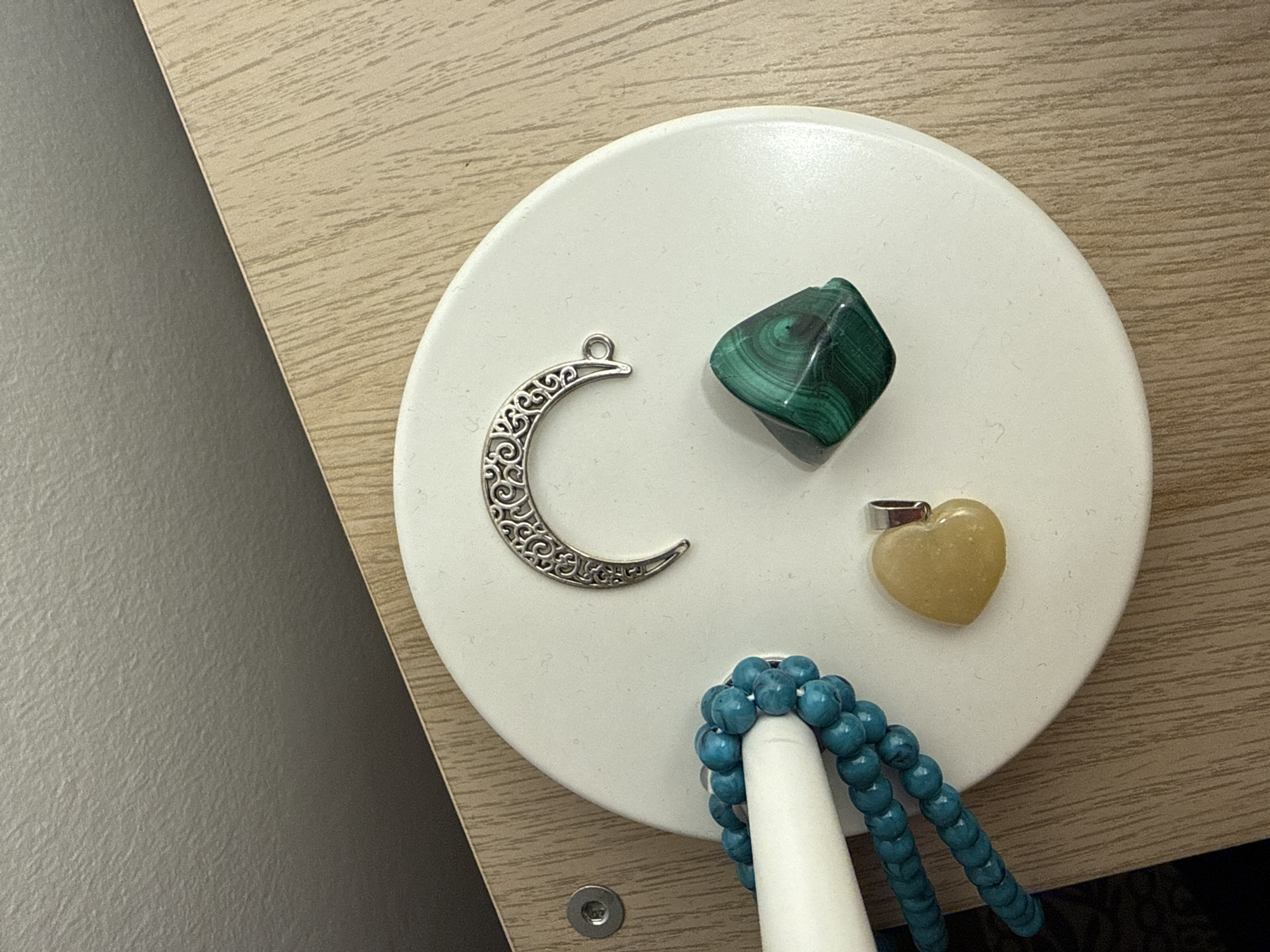 The scene presents a top-down view of various objects arranged on a white, circular surface that appears to be resting on a light-wood-grained surface. Positioned on the white surface are a silver crescent moon charm, a dark green, blocky stone with concentric banding, a pale yellow heart-shaped pendant, and a strand of small, round, turquoise-colored beads. A white, cylindrical object is present near the beads, seemingly supporting or organizing them. The background shows a textured gray wall adjacent to the light-wood-grained surface. There is no evidence to suggest this image was taken in Barcelona, Spain.