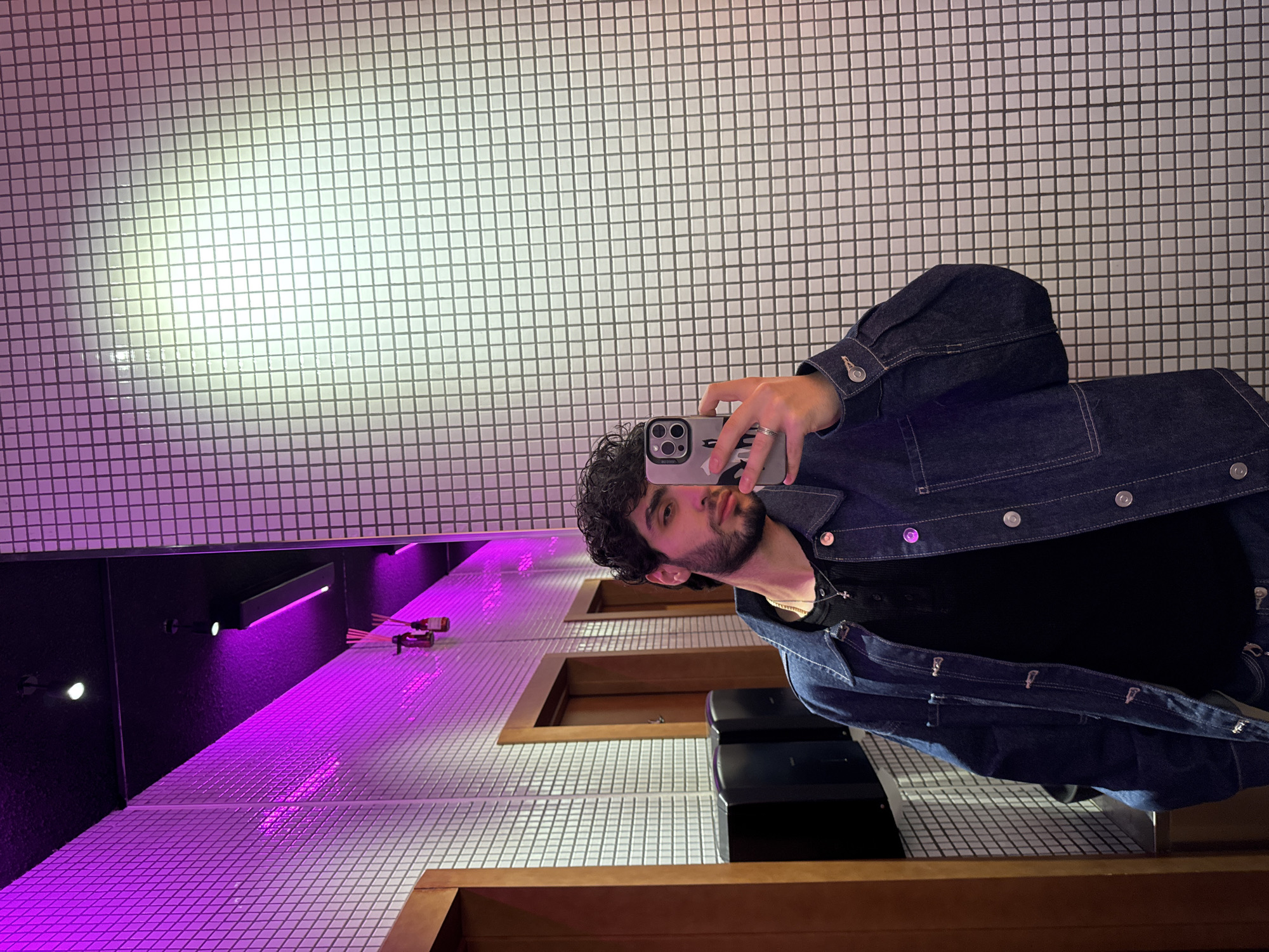 A person is taking a selfie in a restroom with a black and white tiled wall and floor. The restroom features purple lighting. The location is likely in Yerevan, Armenia. The person has curly hair, a beard, and is wearing a dark blue denim jacket over a black shirt. They are holding an iPhone in their right hand. A wooden shelf or panel is visible at the bottom of the frame. There are black rectangular objects on the floor.