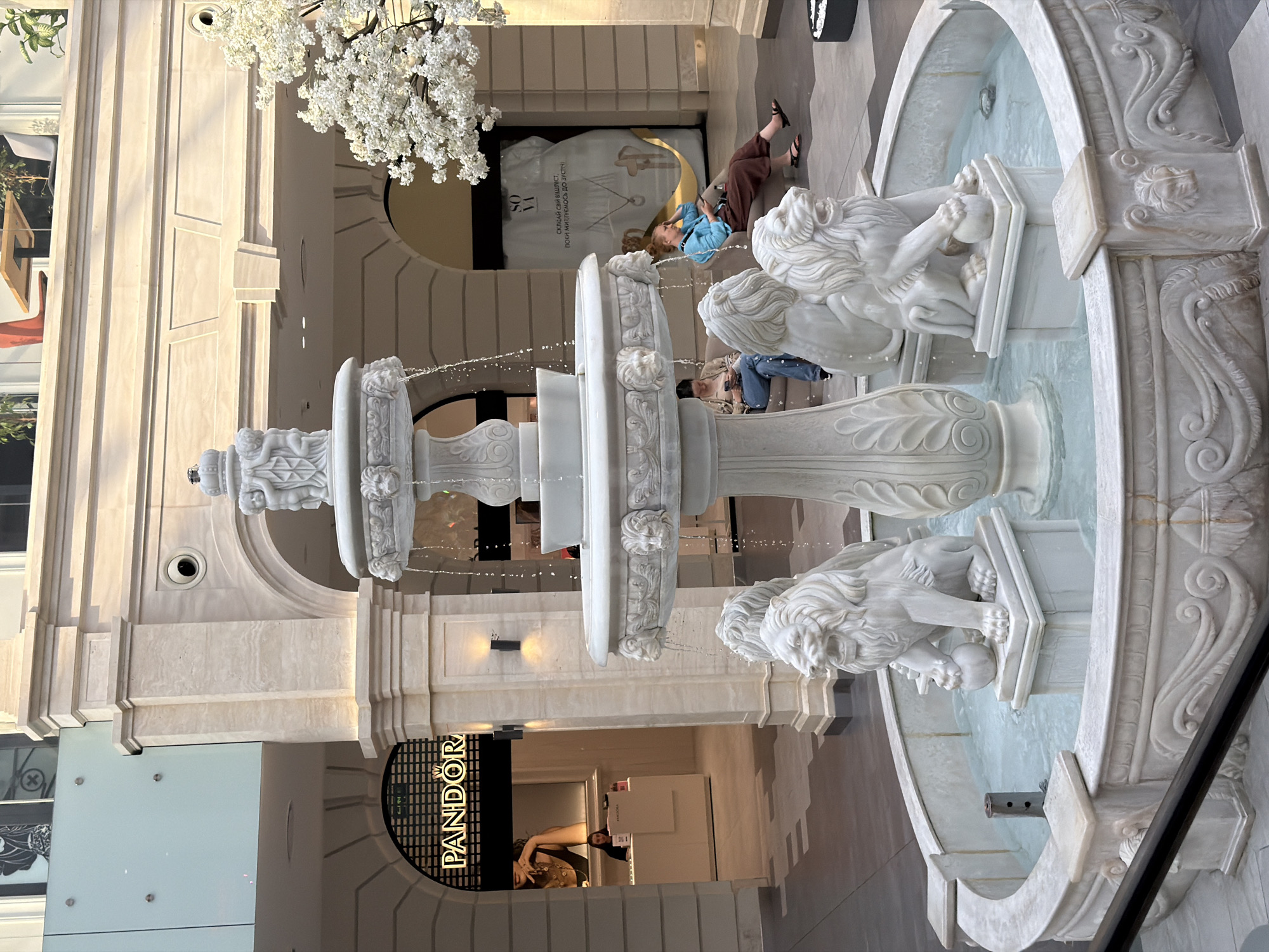 The image shows an indoor fountain with lion sculptures at its base, located in the city of Kyiv, Ukraine. The fountain is made of white marble and features multiple tiers with cherub sculptures at the top. Water cascades down from the upper tiers into the basin. Two women are visible sitting on a bench near the fountain. One woman, wearing a blue top and brown pants, is looking at her phone. The other woman, wearing a beige top, is also looking at her phone. In the background, there is a store with a "PANDORA" sign, indicating it is a Pandora jewelry store. The architecture suggests a modern shopping mall or an indoor public space with classical decorative elements.