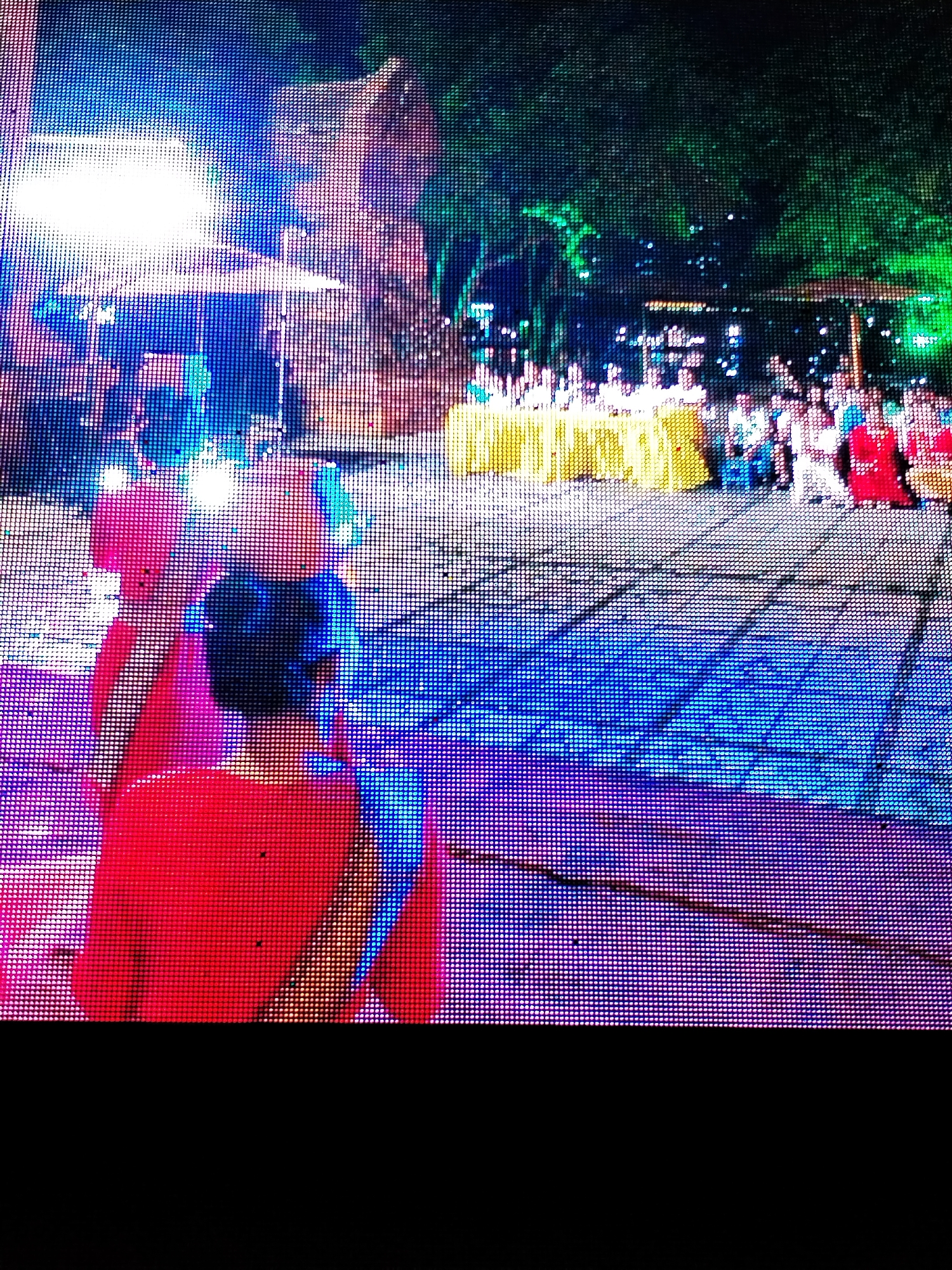 A nighttime scene in Bac Nha Trang Ward, Vietnam appears to be captured on a digital display, showing an outdoor event. In the foreground, a person in a red outfit is visible from the back. Further back, a row of yellow-covered tables stretches across the scene, with people standing and sitting behind them. Trees are visible in the background, illuminated with green light. There are other sources of light, including an intensely bright source that obscures detail behind it, and street lights. The ground is patterned with blue and purple light.