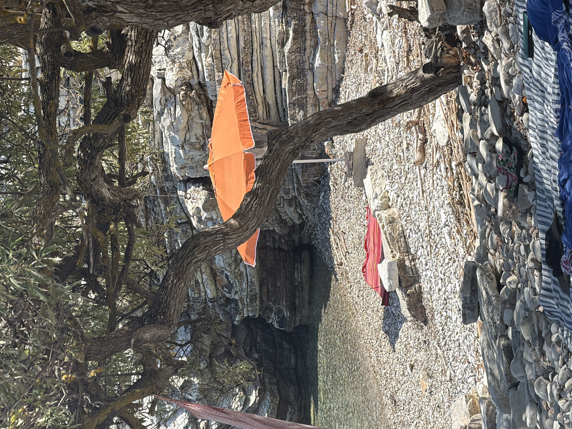 The image depicts a tranquil beach scene in the Municipal Unit of Kastos, Greece, characterized by its natural beauty and rustic charm. A large, gnarled tree dominates the foreground, its branches extending to shade the pebbled shore. Two orange beach umbrellas are visible, one partially obscured by the tree, offering a splash of vibrant color against the muted tones of the landscape.
The beach itself is composed of small, smooth pebbles, leading down to the clear, shallow water of the Aegean Sea. The water's transparency allows a view of the seabed, which is also covered in pebbles. In the background, layered rock formations rise up, creating a natural cliff face with an opening that suggests a small cave or overhang. The rocks are a pale, weathered gray, adding to the rugged aesthetic of the location.
Several items are spread out on the beach, indicating recent human presence. A red and white striped mat or towel is laid out on the pebbles, near a stack of rocks. Further down, a blue and white striped blanket is visible, adorned with a few other items, including a green case and a black sunhat. The presence of these personal belongings suggests that individuals are enjoying a day of relaxation by the sea. The overall atmosphere is one of peaceful seclusion and natural serenity.