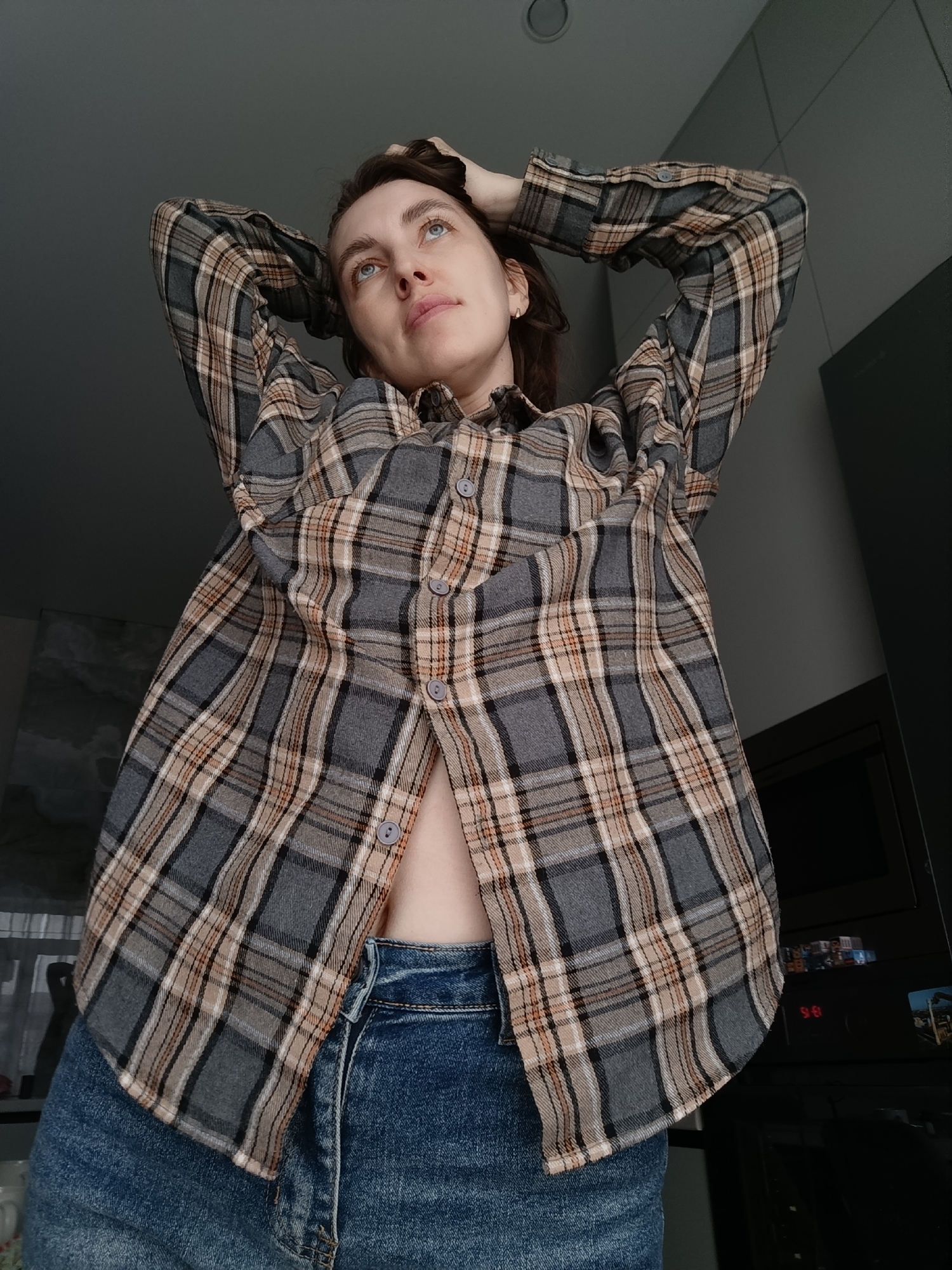 A young woman with brown hair and blue eyes is looking upwards. She is wearing a plaid shirt that is partly unbuttoned, revealing a small part of her abdomen, and blue jeans. Her hands are raised and holding the back of her head. The background includes a ceiling, a dark-colored cabinet, and a window. There is no definitive evidence that the image was taken in Chaikovsky, Russia.