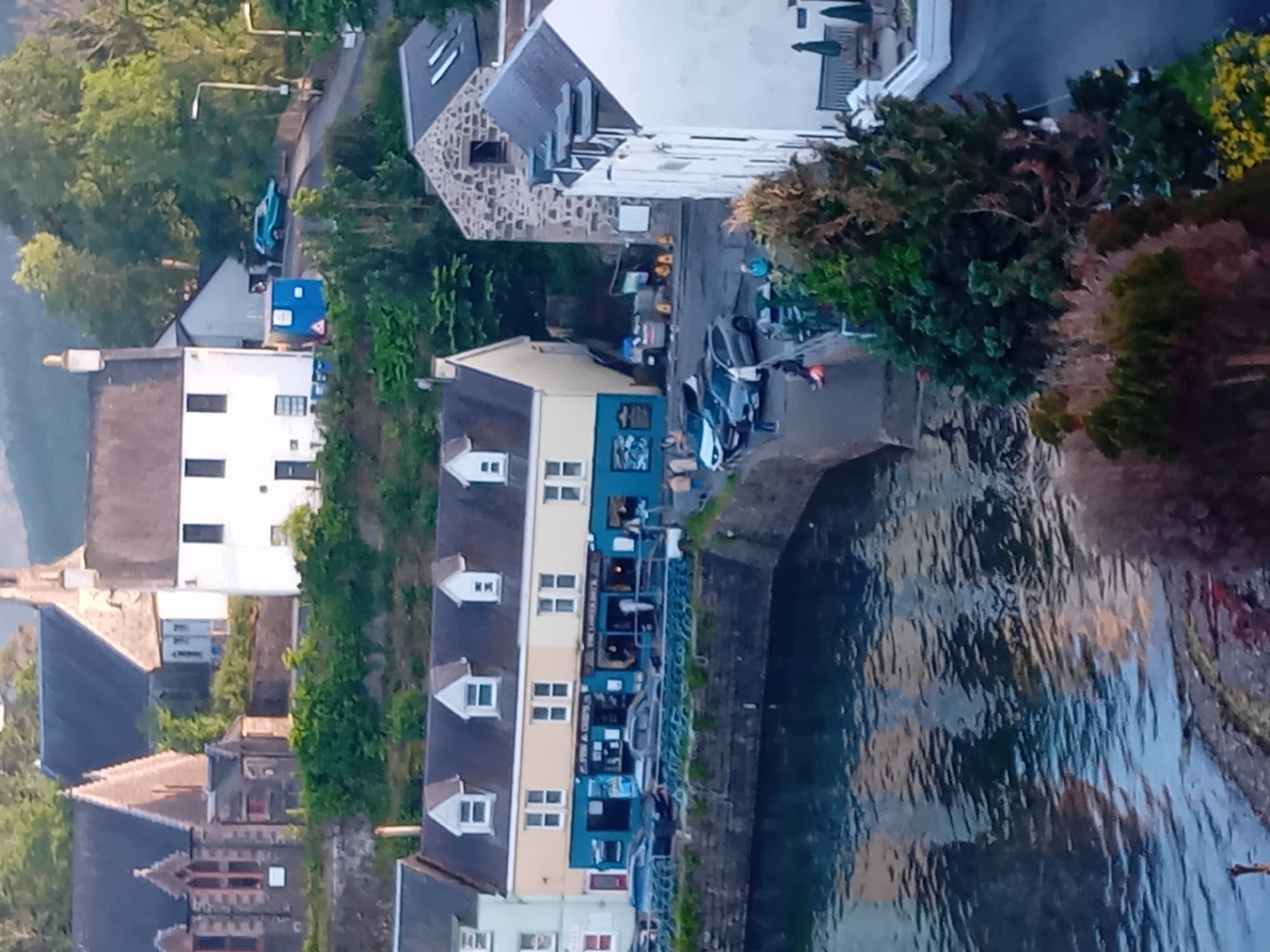 This is an eye-level, medium shot of a waterfront in Portree, United Kingdom, with buildings lining the harbor. In the foreground, the calm, blue water reflects the colors of the buildings and sky. A stone retaining wall runs along the edge of the water, and various types of vegetation, including trees and bushes, are visible.
Several buildings are situated along the waterfront. One prominent building is painted yellow and blue, with multiple windows and dormers. Below this building, a street or walkway runs along the harbor, with several parked cars. A sign on the yellow and blue building appears to read "FISH & CHIPS" and "THE LOCHINVAR INN." Another building, painted white with dark windows, is visible to the right of the yellow and blue structure. Further in the background, more buildings, including what appears to be a church with a tall steeple, are nestled into a hillside covered in lush green trees. A light blue vehicle is parked on a street in the upper right portion of the image, near a lamppost. The sky is overcast and a soft light illuminates the scene.