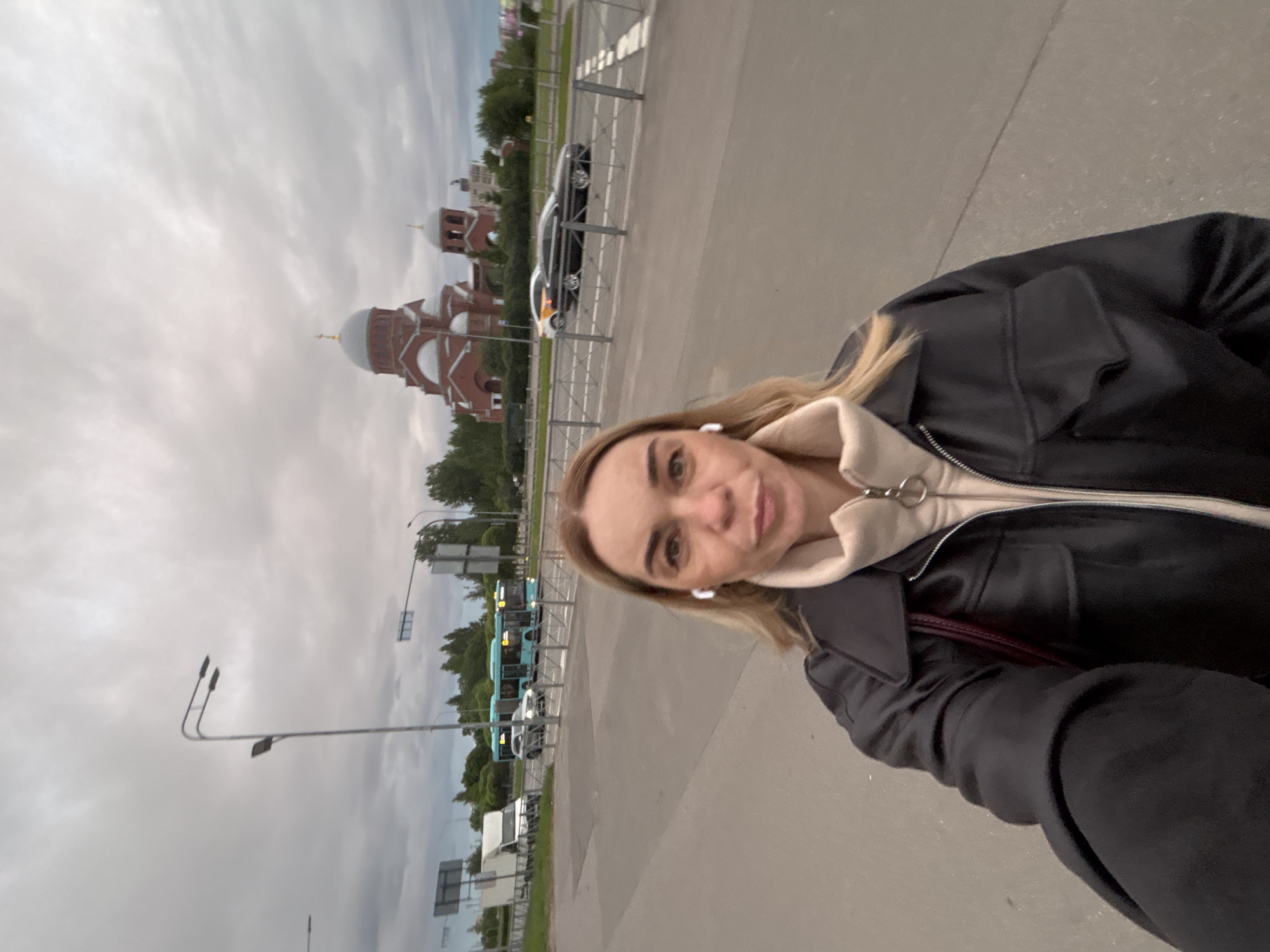 The image shows a woman standing on a sidewalk next to a road. In the background, there is a church with a large dome. There are also cars and buses parked on the side of the road, as well as streetlights. The sky is overcast.
Given the architectural style of the church, it is plausible that the location is in Russia. It is not possible to determine the city without further information.