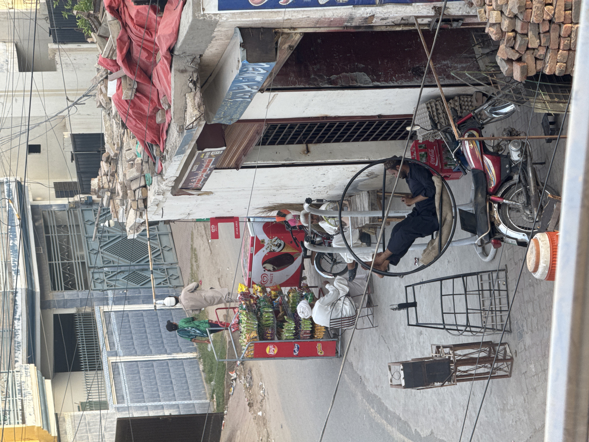 This image captures an outdoor street scene in Unknown, Unknown, under daylight conditions, likely morning or late afternoon, given the soft shadows. The perspective is elevated, looking down onto the street and adjacent structures. In the immediate foreground, a metal railing runs horizontally across the bottom, partially obscuring a white and orange insulated water cooler. Multiple black utility wires crisscross the frame from foreground to midground. The midground features a paved street on the left, transitioning to a dirt path towards the background. Two men are walking away from the viewer on the left side of the street. The man closer to the camera wears a green and blue patterned shirt and dark trousers. The man in front wears a light beige tunic (shalwar kameez) and has a white object, possibly a small container or cloth, balanced on his head. On the right side of the street, a small commercial setup is visible. A red metal stand displays various packaged snacks, including Lay's, Kurkure, and Cheetos brands. Adjacent to this is a red and white Wall's ice cream freezer, depicting an ice cream cone and a heart design. A man in a white turban and white tunic is seated on a brown metal chair next to the snack stand, facing right. Further right, another man in dark clothing is reclined in a circular metal swinging chair, holding a mobile phone to his right ear. His feet are elevated and propped up. A red Honda CD 70 motorcycle is parked nearby, facing right. Next to the motorcycle, stacks of brown egg cartons and red bricks are visible against a building facade. Two empty metal frame structures, one appearing to be a stand or cart, are positioned on the pavement. The background consists of several multi-story buildings. On the left, a building features a blue-tiled lower facade with a black wrought-iron fence and a large, ornate metal gate. Other buildings exhibit varying states of finish, from smooth stucco to exposed concrete. On the right, a dilapidated building has two dark doorways; one is covered by a metal grate, and the other is open, revealing a dark interior. Signs with text in an unknown script are mounted above these doorways, one with blue text on a white background. The roof of this building and an adjacent structure are covered with debris, including loose bricks, concrete blocks, and a large red tarp draped over