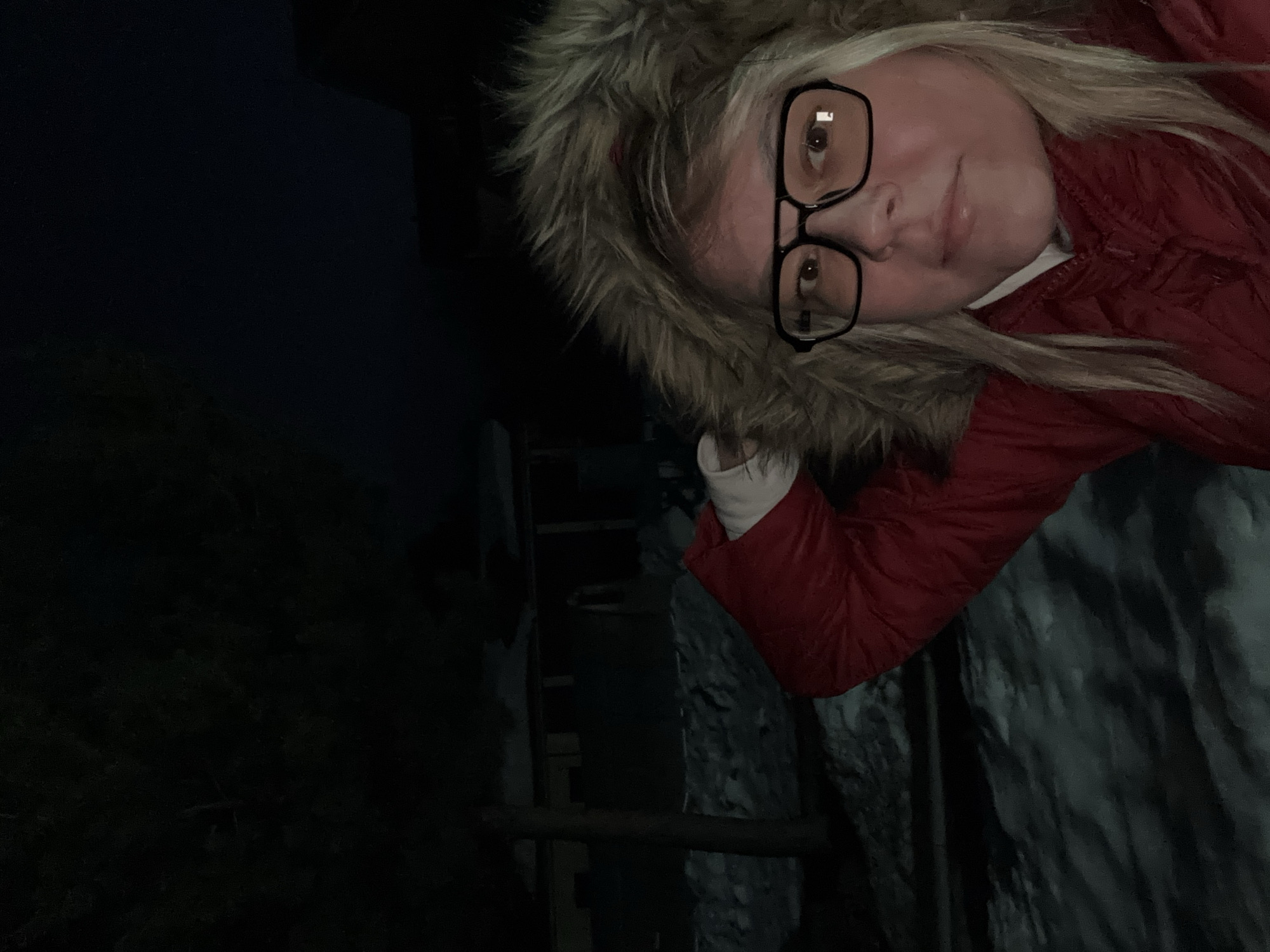 The image depicts a female individual in a snowy outdoor environment at night. The subject is a woman with blonde hair, wearing black-framed glasses, a red quilted winter jacket with a light-colored fur-lined hood, and a white garment visible at the neck. Her right arm is raised, with her hand positioned near the top of her head, suggesting a self-portrait. She is looking directly towards the camera. The ground in the foreground and midground is covered in snow, indicating winter conditions. In the dark background, indistinct outlines of trees and low-rise structures are discernible. The scene is located in the city of Русский Вожой, Russia.