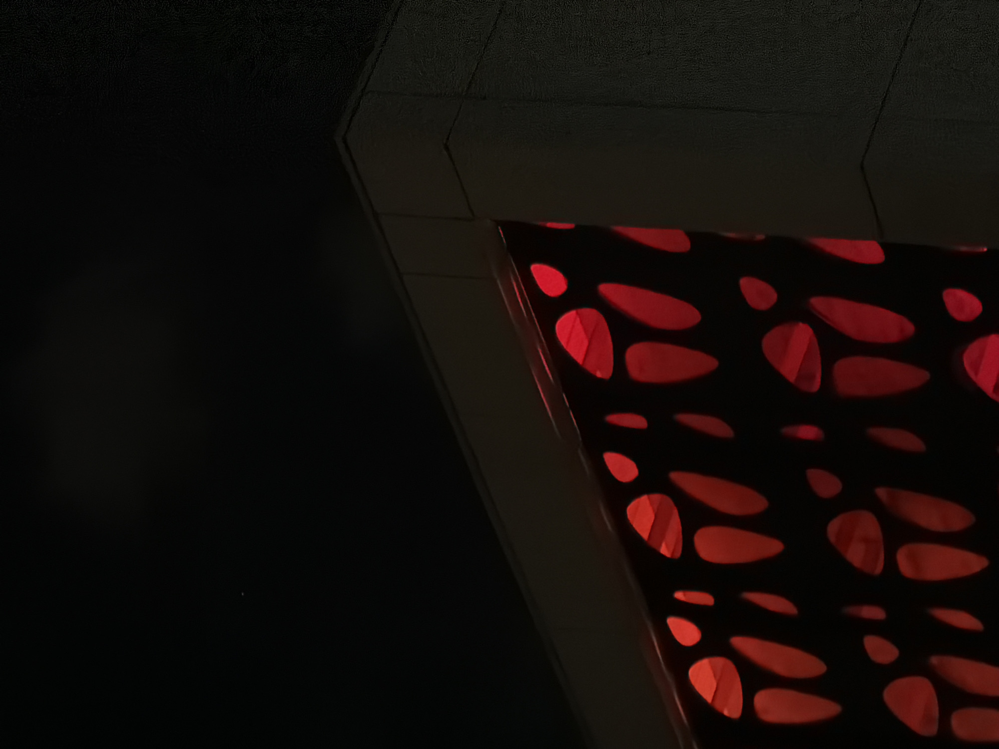 A dimly lit, close-up shot captures a section of an architectural feature in Tel-Aviv, Israel. The scene is dominated by a dark surface, possibly a ceiling or wall, with a textured appearance. A lighter-colored, angled structure juts out, leading to a decorative panel adorned with red, organic shapes, like stylized leaves or petals. The shapes are illuminated, creating a striking contrast against the dark background. No people are visible.