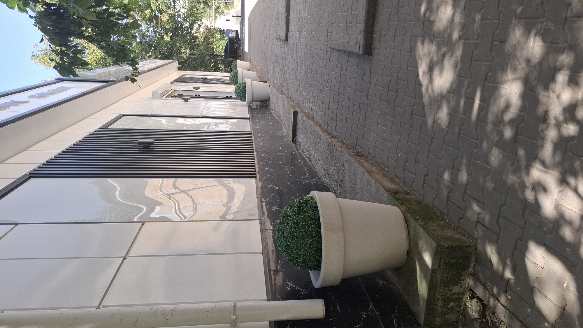This image depicts the exterior of a modern building located in Shymkent, Kazakhstan. The building features a facade with white tiled panels and reflective windows, interspersed with vertical dark slats. The ground level is bordered by a dark, marble-patterned lower section. Along the sidewalk, several large, light-colored planters contain round, green artificial topiary plants. The sidewalk is paved with interlocking gray bricks, and dappled sunlight creates shadows from overhead trees. A white downspout is visible on the left side of the building. In the background, trees and a distant parked vehicle suggest an urban setting. The overall impression is one of a clean, well-maintained commercial or public space.