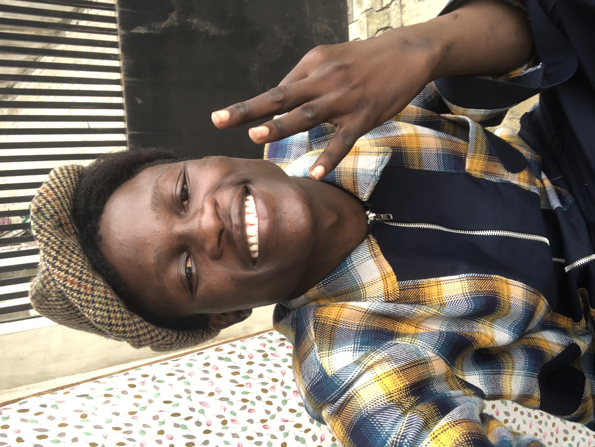 A young Black male wearing a plaid jacket and a flat cap is posing for a photo, holding up two fingers on his right hand. He is smiling and looking directly at the camera. The background appears to be outdoors, with a metal fence and a patterned curtain visible. The image was taken in Ado, Nigeria.