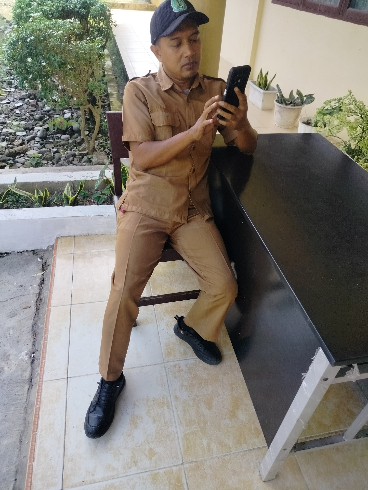 A man is seated in a chair outside, next to a black table, looking at his phone. He is wearing a tan uniform, a black baseball cap, and black sneakers. The environment appears to be a courtyard with tiled flooring and potted plants. Rocks and vegetation are visible in the background. The location is identified as Lawebekung, Indonesia.