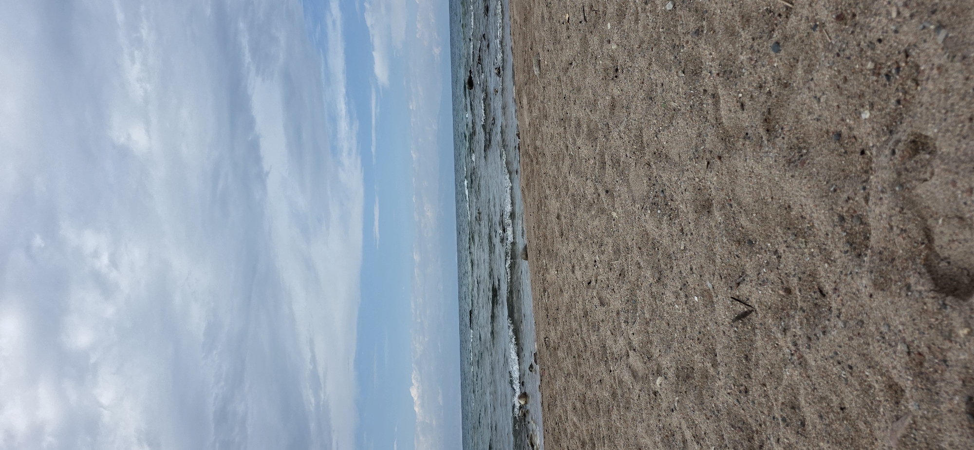The image depicts a beach scene with a sandy foreground leading to the water. The water is calm with small waves lapping at the shore. The sky is overcast with scattered clouds, suggesting a potentially cool or breezy day. In the background, a distant landmass is visible, likely mountains or hills bordering the water. There are no people or animals visible in the frame. The image was taken on a beach in the city of Tong, Kyrgyzstan.