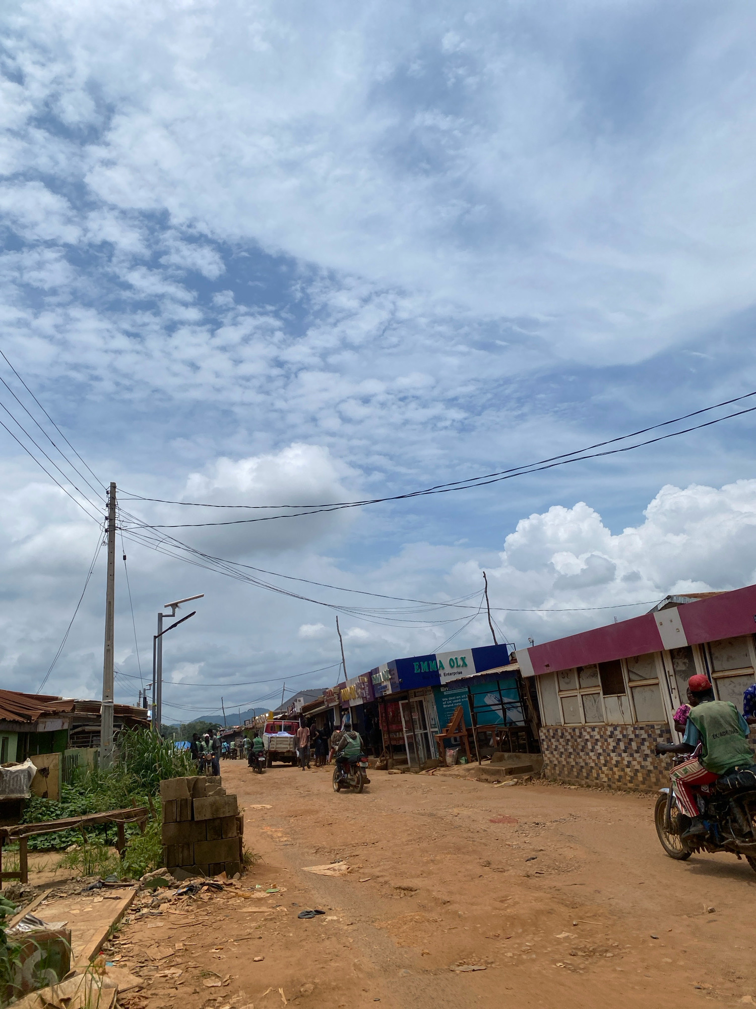The image depicts a street scene in Iworoko, Nigeria. The street is unpaved and appears dusty, with a slight incline. On both sides of the street are buildings that house various businesses, some with corrugated metal roofs and others with more modern facades. Signs for "EMMA OLX" and "Enterprise" are visible on one building. Electrical wires are strung across utility poles, and a solar-powered street lamp is present. In the foreground, stacks of concrete blocks and debris litter the ground. Several people are present, with some riding motorcycles and others walking. A truck is parked further down the street, and various goods are displayed outside some of the shops. The sky is partly cloudy with significant cumulus formations. The overall impression is of a busy, somewhat rustic marketplace in a Nigerian town.