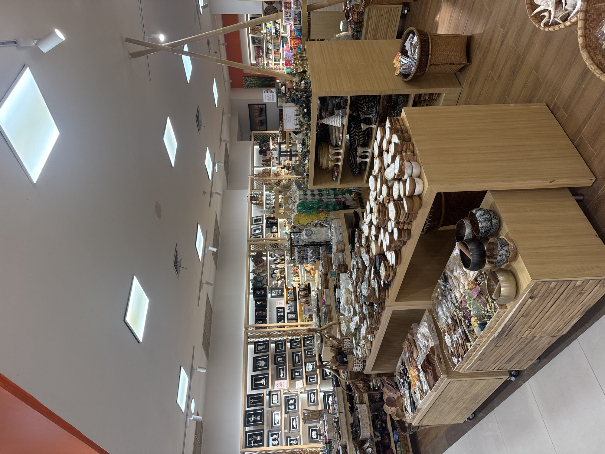 The image shows the interior of a well-lit store selling handcrafted items and home decor, likely located in Kuta Selatan, Indonesia. The store features wooden display shelves filled with a variety of products, including framed art, ceramic bowls, wooden utensils, and decorative figurines. There are several wooden tables in the foreground showcasing stacks of bowls and utensils. Natural light streams in from ceiling panels, supplemented by track lighting. The overall atmosphere is one of a boutique market or souvenir shop. There are no people visible in the scene.