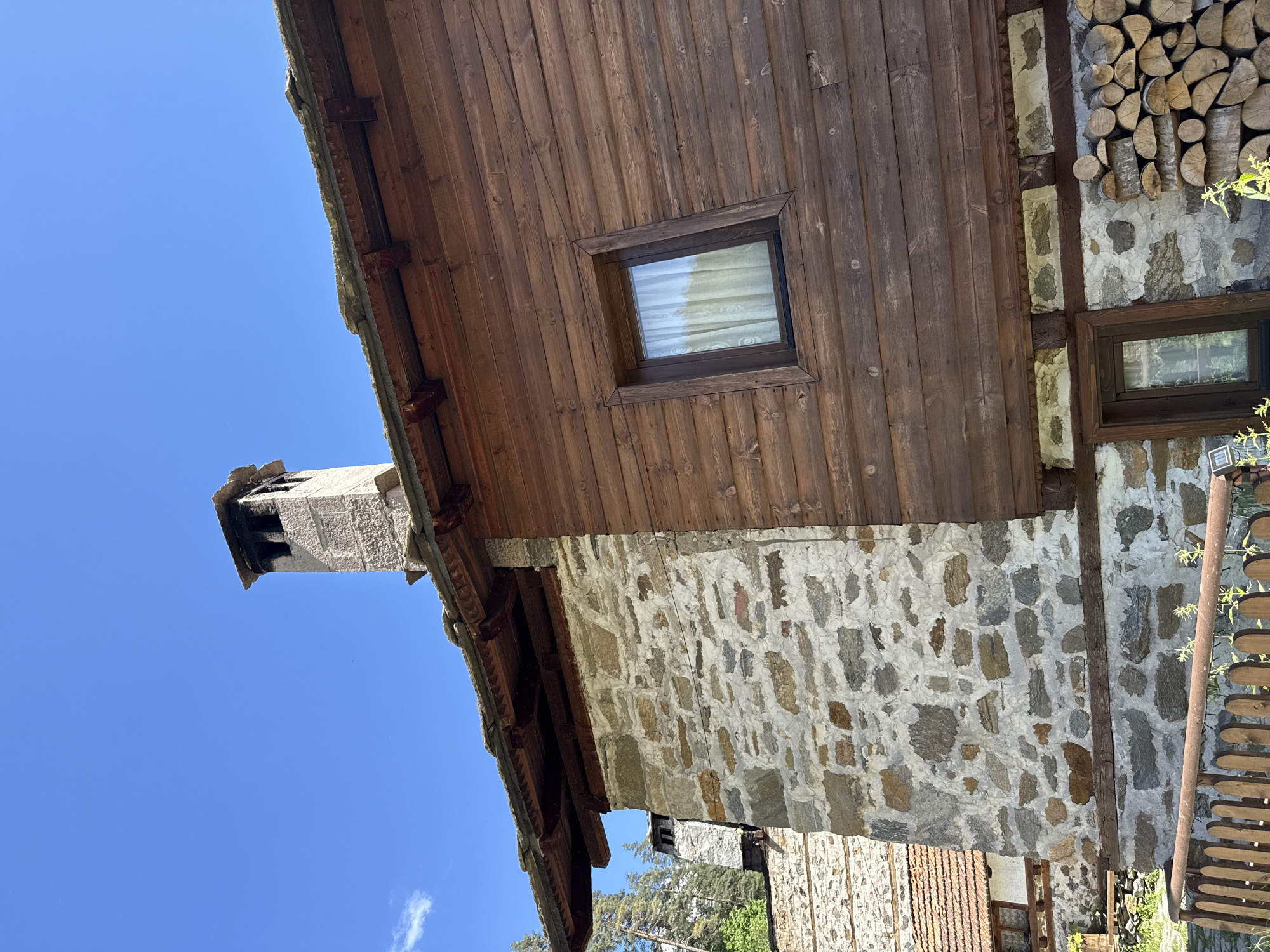 The image shows a traditional Bulgarian house in the village of Shiroka Laka. The house has a stone foundation with wooden upper walls. The roof is made of stone slabs. There is a chimney made of stone on the roof. A window with white curtains is visible on the wooden wall. In front of the house, there is a pile of firewood. The sky is clear and blue. The overall impression is of a rustic and charming rural dwelling.