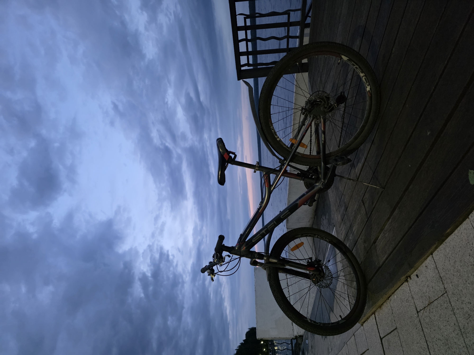 The image shows a black and red Stels mountain bike lying on its side on a wooden surface, likely a boardwalk or deck. The sky in the background displays a cloudy, twilight or dusk scene, with hues of blue and some pink/orange near the horizon. Behind the bike, a metal railing suggests the edge of the boardwalk, with water visible beyond. The surface beneath the front wheel of the bike appears to be cobblestone or paving stones. Given that the image has been identified as being located in Chaikovsky, Russia, the water in the background is likely the Kama River which runs through the city of Chaikovsky. The specific location on the riverfront can not be determined from the provided image. No people are visible.