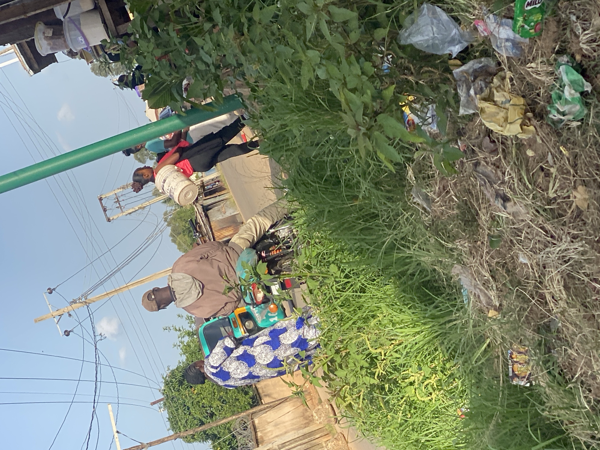 The image depicts an outdoor scene in Jos, Nigeria, likely during the day with clear skies. The perspective is from ground level, looking up at a slightly elevated view of people and their surroundings.
In the foreground, thick green grass and dry weeds are prevalent, with visible litter scattered among them, including plastic bags and wrappers.
Three individuals are visible. One person, wearing a red shirt and black pants, is partially obscured by a large green pole. They are holding a white bucket. To their right, another person, wearing a brown jacket and a cap, is seated on what appears to be a three-wheeled vehicle, possibly a motorcycle or a tricycle common in the region. This person is looking down. In front of the vehicle, a third individual, wearing a blue and white patterned dress, is also seated. The three-wheeled vehicle is turquoise and yellow and has a visible headlight.
In the background, a wooden structure or stall is visible, with several white buckets stacked on it. Overhead, power lines and utility poles are prominent against the blue sky. The overall scene suggests a public space, possibly a roadside or market area, where daily activities are taking place amidst signs of neglect due to litter. The presence of the power lines and the type of vehicle hint at a semi-urban or rural setting within Jos. No discernible text is clearly legible in the image.
