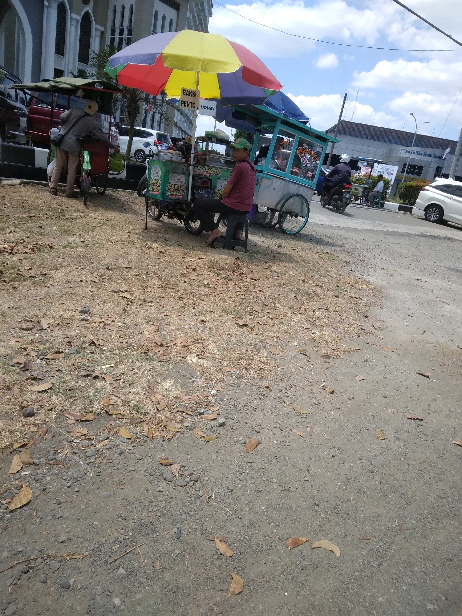 In a sun-drenched outdoor scene on a dirt and gravel lot next to a paved road, a food cart with a colorful umbrella is the central focus. The cart, painted green and silver with blue trim, is laden with goods and appears to be selling food items, with visible signage indicating "BAKSO PENTOL" and "SIOMAY". A person wearing a maroon shirt and a green cap sits on a small stool next to the cart, seemingly attending to it. To the left of this cart, another food cart, red and green, is partially visible, with a person in beige attire and a hat standing near it, possibly interacting with the cart or its contents. In the background, a large, imposing building with arched windows stands prominently, its facade suggesting a public institution or place of worship. Several vehicles are also present: a red SUV, a white car, and a motorcycle with a rider wearing a white helmet. The ground is covered with dry leaves and small stones, indicating a possibly arid or autumnal environment. The sky above is a clear blue with scattered white clouds. This scene is located in Sokaraja, Indonesia, as indicated by the context.