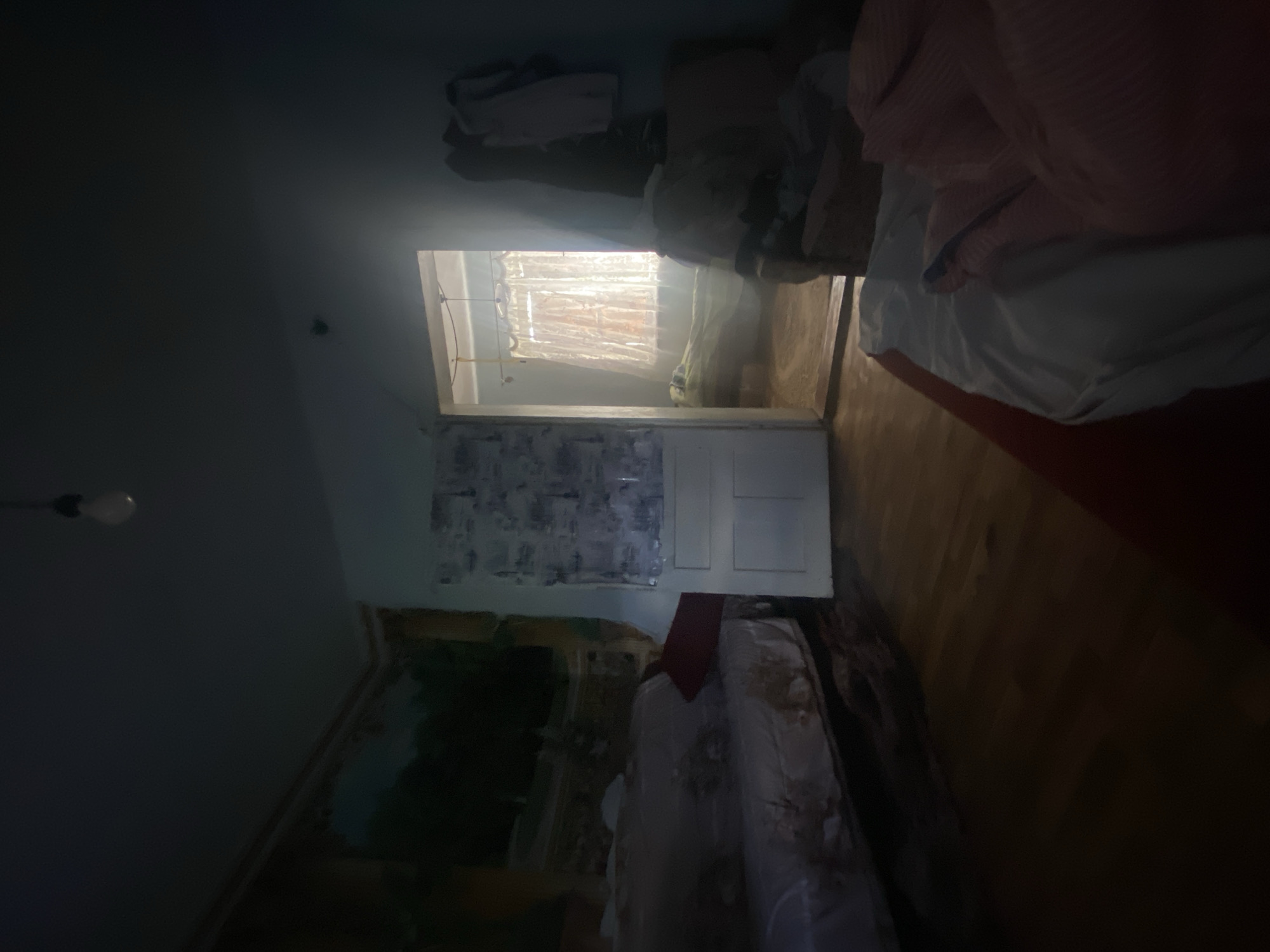 The image depicts a dimly lit bedroom in a dwelling located in Borshchiv, Ukraine. The room appears to be in a state of disarray. In the foreground, a bed is visible with rumpled white and pink bedding. To the left, another bed or sofa is partially covered with a patterned fabric, and a red pillow is propped up against it. A large tapestry with a landscape scene hangs on the wall to the left. A doorway in the center of the image leads to an adjacent room where a window with sheer curtains is visible, allowing light to filter through. Clothes are hanging on the right side of the doorway. The floor is made of wooden planks. A bare light bulb hangs from the ceiling. The overall atmosphere is dark and somewhat neglected.
