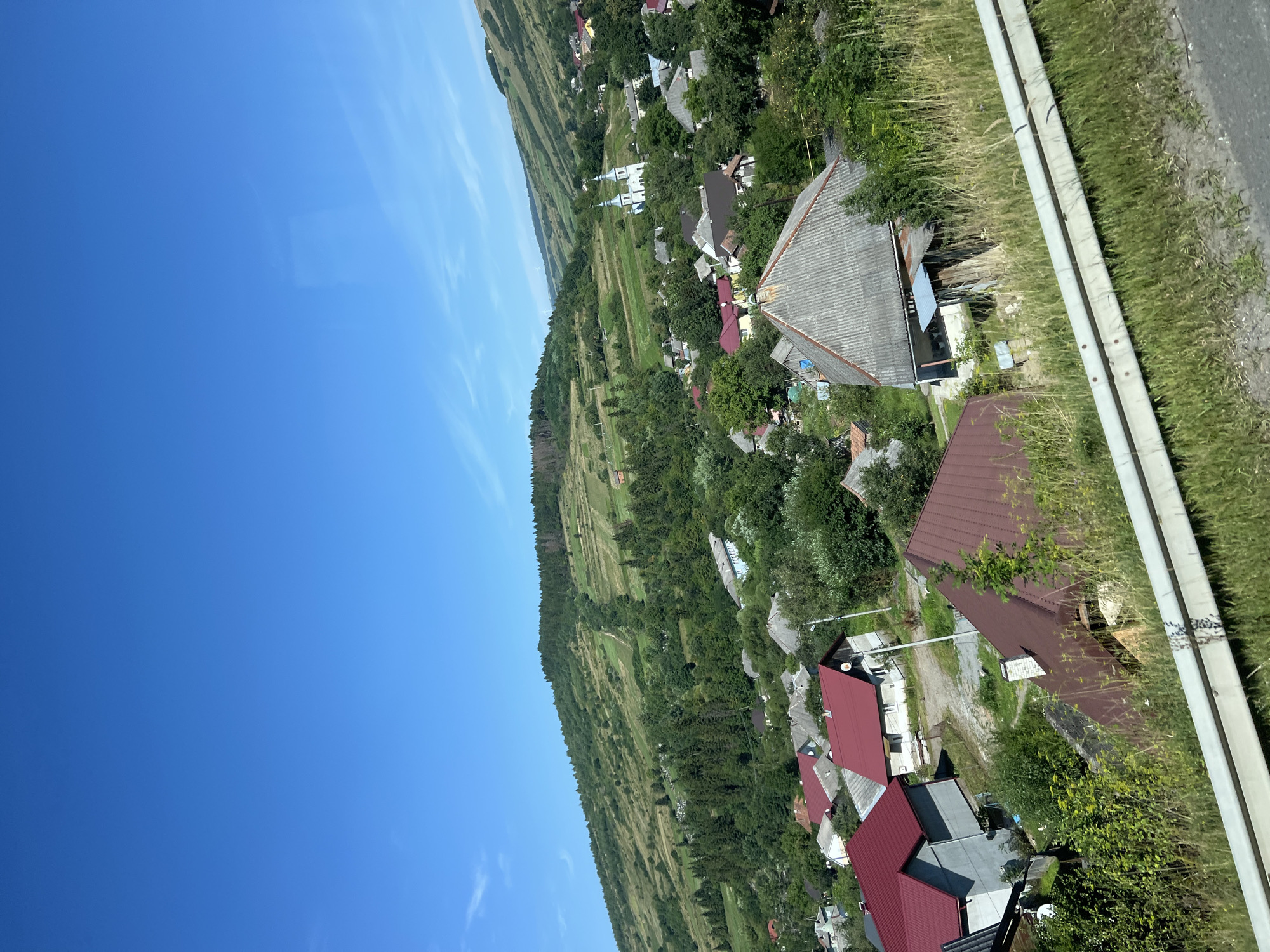 The image captures a view of the village of Nyzhni Vorota, Ukraine, nestled within a valley and surrounded by lush green hills. The sky is a clear, vibrant blue, with a few wispy clouds. In the foreground, a white guardrail runs horizontally across the bottom of the frame, with tall, dry grass growing on either side.

The village itself is a collection of houses with various roof colors, including red and gray, scattered amongst dense green trees. Several buildings are visible, some appearing to be residential homes. In the mid-ground, a prominent church with twin white spires stands out against the backdrop of the village and the rolling hills. The hills are covered in a mix of evergreen trees and cultivated fields, showing evidence of human activity and agricultural practices. The overall impression is a picturesque rural landscape bathed in bright daylight. There are no people visible in the image.