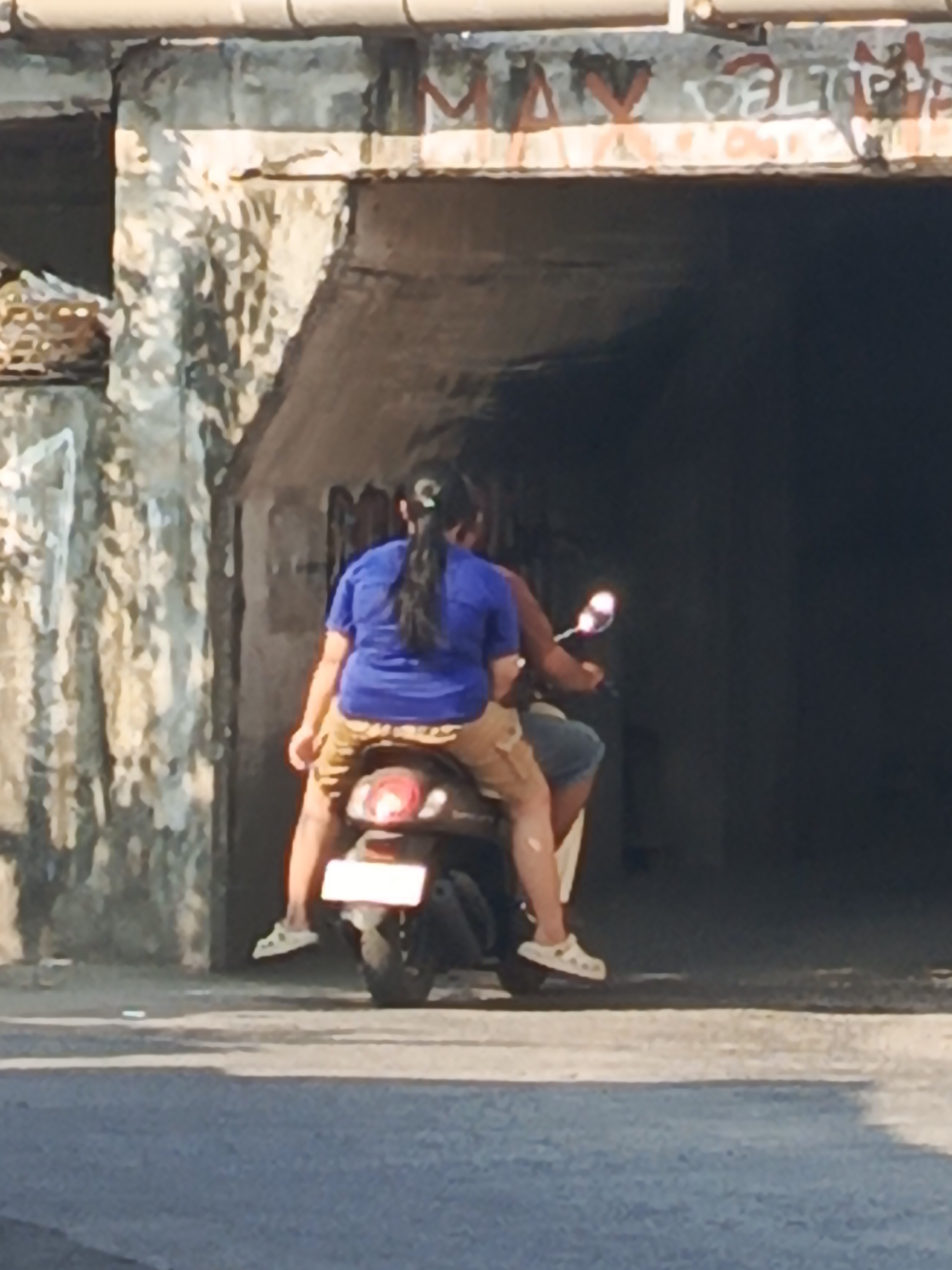 The image depicts two individuals on a scooter beneath a concrete overpass with graffiti. The person in the rear, wearing a blue t-shirt and patterned shorts, is operating the scooter. The person in the front, whose clothing is obscured by the rider, is facing away from the camera. The scooter has a white license plate and a visible taillight. The environment appears to be an urban setting with a paved road in the foreground and textured concrete walls on either side of the overpass. Graffiti is visible on the underside of the overpass. The scene is lit by sunlight, casting shadows and highlighting the textures of the concrete structures. This scene is located in Kludan, Indonesia.