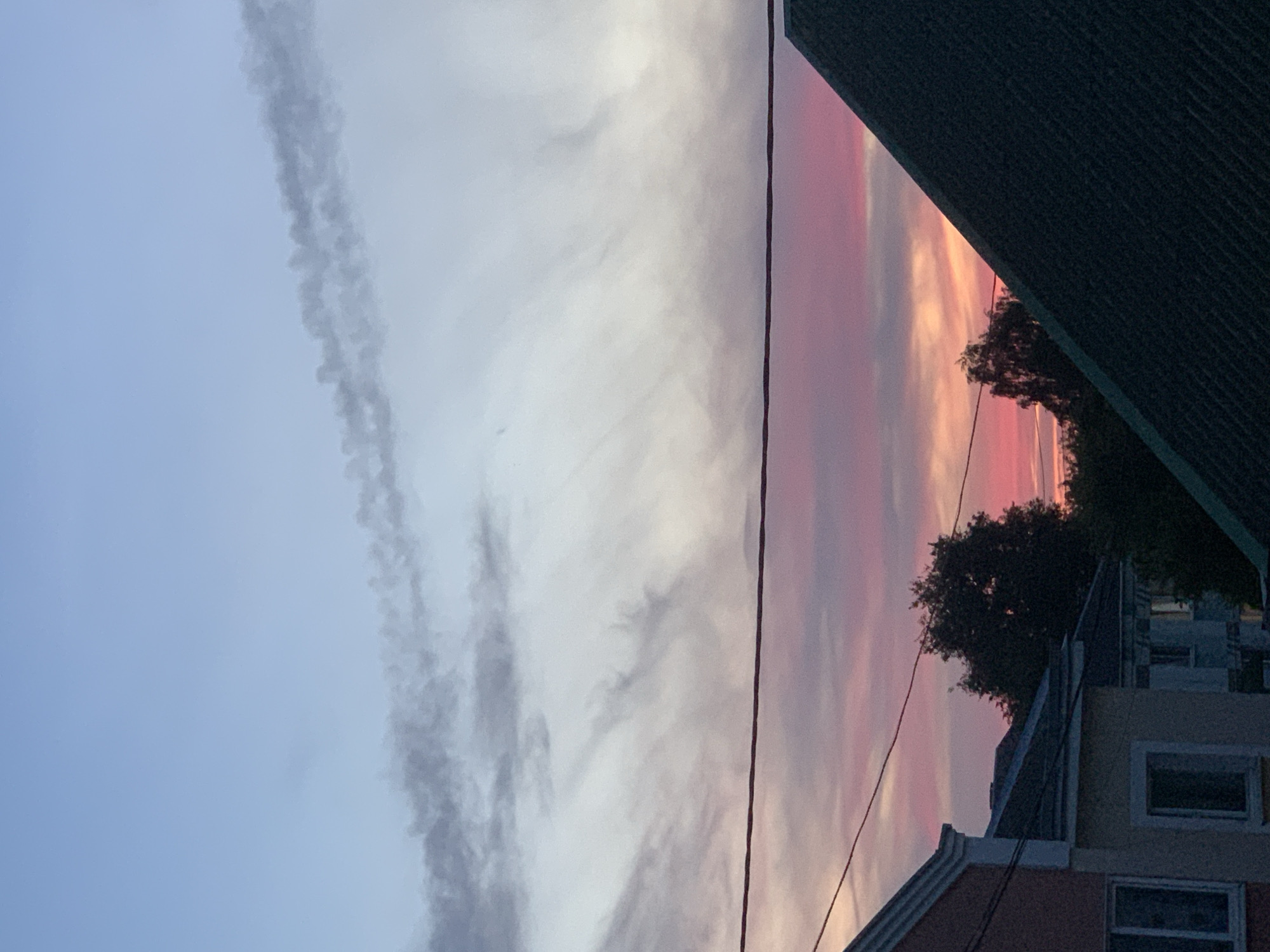 The image depicts a sunset sky over a residential area in the city of Kapyĺ, Belarus. The sky transitions from a light blue at the top to vibrant pink and orange hues near the horizon, with wispy clouds scattered across. A dark horizontal line, likely a power line, runs across the mid-section of the frame. In the foreground, the roofs and upper portions of houses are visible, characterized by their sloped structures and muted colors. Silhouetted trees are present behind the houses, and a prominent dark, triangular roof dominates the lower right portion of the image. The overall scene captures a calm, twilight atmosphere.