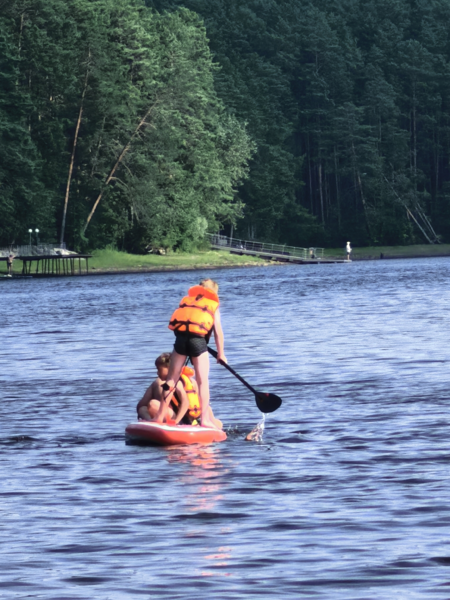 In this image, two individuals are paddleboarding on a calm body of water, possibly a lake, as indicated by the presence of a distant wooden pier and a tree-lined shore. A dense forest forms the backdrop, suggesting a natural, possibly secluded, environment. The water is a deep blue, reflecting the clear sky, and small ripples suggest gentle movement. The scene appears to be set during the daytime, with soft lighting that hints at either early morning or late afternoon.
The two people on the paddleboard are wearing bright orange life jackets, prioritizing safety. One person, standing, is actively paddling with a black paddle, creating a splash of water as they move. The other person is seated on the front of the board. Both individuals appear to be enjoying the activity, with no overt signs of distress. The paddleboard itself is a bright red and white inflatable.
In the background, a wooden pier with multiple supports extends into the water, and several people can be seen on or near it. Further in the distance, more people are visible, some appearing to be walking along the shore or on another platform. The overall atmosphere is serene and suggests a recreational outing in a natural setting. The location is identified as Sysert, Russia. There is no visible text in the image.