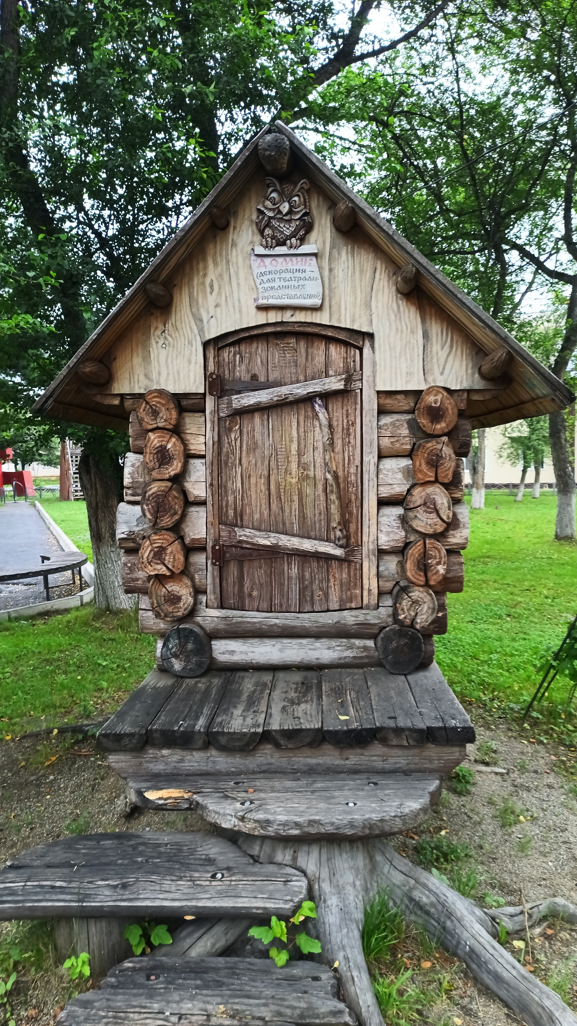 The image shows a small wooden structure resembling a miniature log cabin, set within a park or garden area. The structure features a pitched roof, a rustic wooden door with cross bracing, and is built from logs of varying diameters. Above the door, there is a decorative carving of an owl and a plaque with Cyrillic text. The structure stands on a platform made of rough-hewn wooden planks and logs. The surrounding environment consists of green grass and trees. The overall impression is one of a whimsical, rustic, and possibly decorative outdoor feature. The structure is located in the city of Abaza, Russia, and appears to be a part of the "Forest Fairy Tale" park.