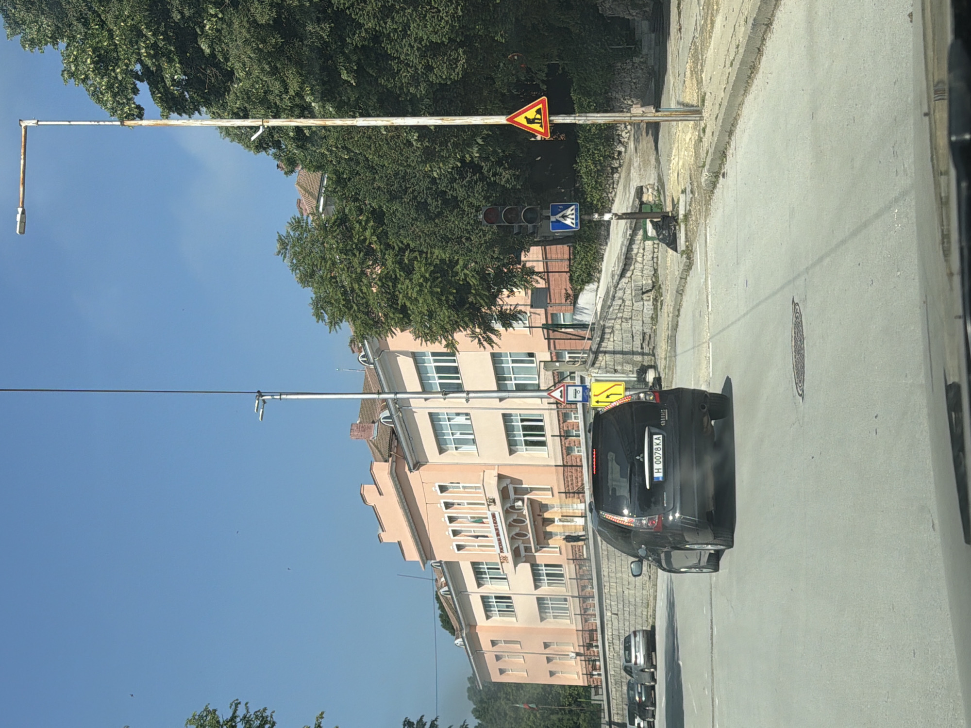The image captures an urban street scene in Shumen, Bulgaria, on a clear, sunny day with a bright blue sky. A black Mitsubishi car, bearing the Bulgarian license plate "H 0078 KA," is visible on the asphalt road in the foreground, seemingly stopped or parked.
To the left, a prominent multi-story building with a light peach or pink facade dominates the background. This building features multiple windows, an ornate entrance, and possibly flags or banners, with partially obscured text on a sign above the entrance. Lush green trees line the street, partially obscuring other buildings and the sky.
Several street objects are present:
* **Traffic Signals:** A set of vertical traffic lights with visible red, amber, and green lenses.
* **Street Signs:** A triangular yellow warning sign with a black border, depicting a person working (likely indicating road work or construction ahead). Below it, a blue square sign with a white pedestrian symbol marks a pedestrian crossing. Further down the street, another yellow diamond-shaped sign with black arrows (possibly indicating a winding road or double bend) is visible.
* **Streetlight:** A long, rusty white utility pole with an L-shaped arm holds a streetlight.
* **Infrastructure:** A concrete curb separates the road from a cobblestone sidewalk. A manhole cover is visible on the asphalt.
No people are clearly visible, but the pedestrian crossing sign suggests foot traffic. The scene depicts a typical European city street environment during daylight hours.