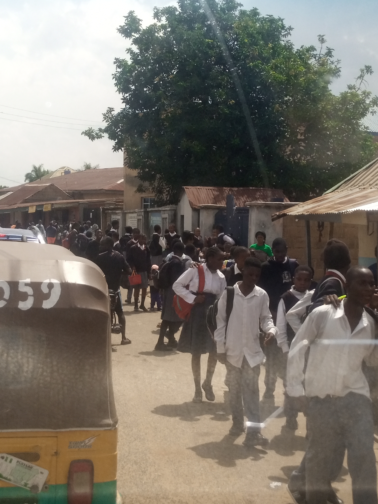 The image depicts a crowded street scene with a large group of young people, likely students, dressed in school uniforms. They are predominantly wearing white shirts, dark skirts or trousers, and some have blazers. Many are carrying backpacks, and some are holding other bags. The individuals appear to be walking in various directions, suggesting a transition, perhaps from school or to a school event.
The setting is an outdoor urban environment. Buildings with corrugated metal roofs and what appear to be shops line the street. There is a large tree providing shade, and overhead power lines are visible. The ground is paved, and shadows indicate it is daytime. The sky is overcast.
In the foreground, a portion of a three-wheeled vehicle, possibly an auto-rickshaw, is visible. It is yellow and green, with a gray canopy. The number "959" is prominently displayed in white on the side of the vehicle. A license plate with some illegible text is attached to the vehicle.
Some of the students are interacting with each other, while others are moving with purpose. Their expressions are not clearly discernible due to the distance and image quality, but there is no indication of distress or unusual activity. The overall impression is one of a busy school day or a period of movement related to school.