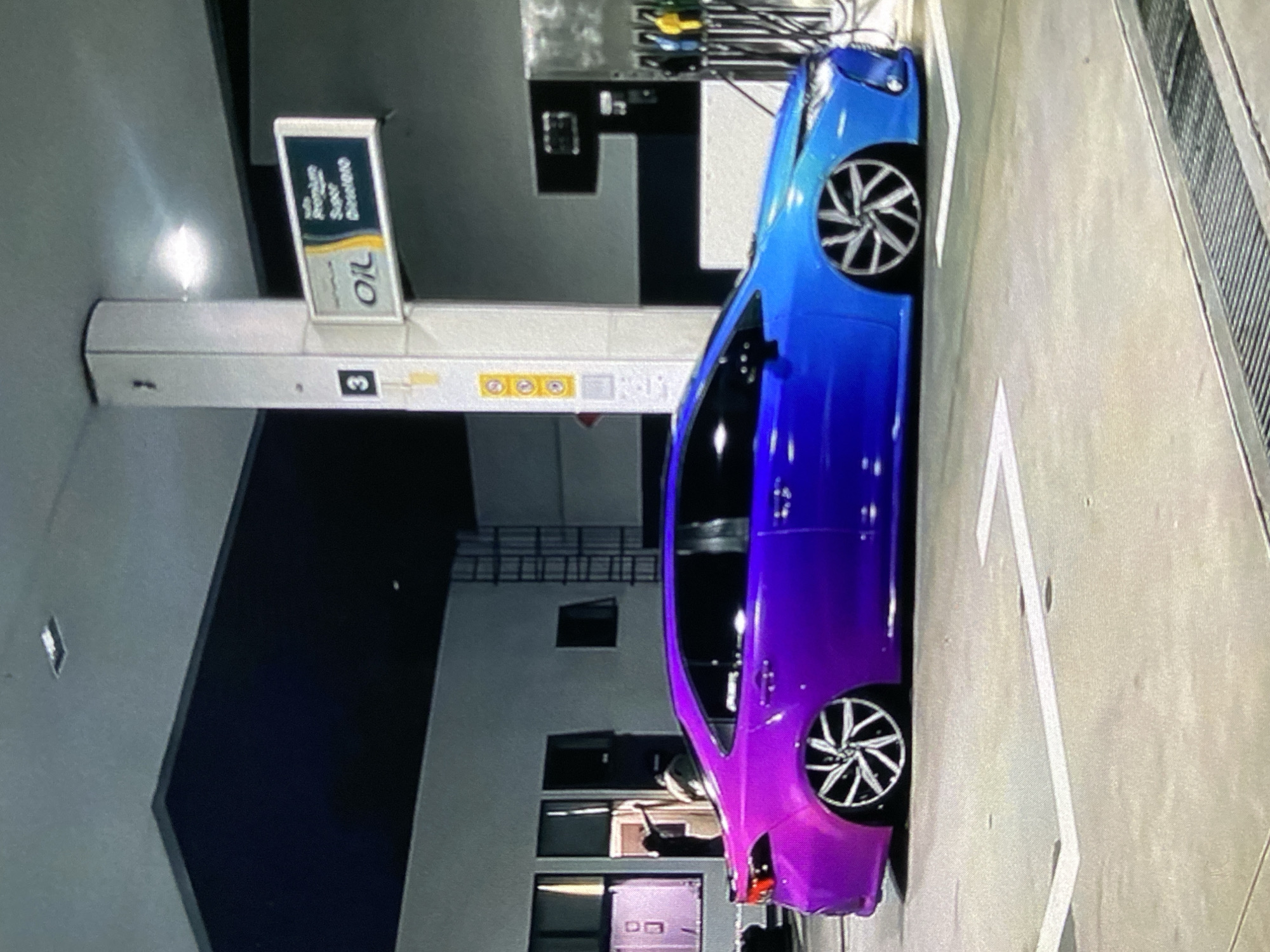A blue and purple car is parked at a petrol station. The petrol station has signs for "Oil" and "Premium Super diesel". The location is Lhokseumawe, Indonesia.