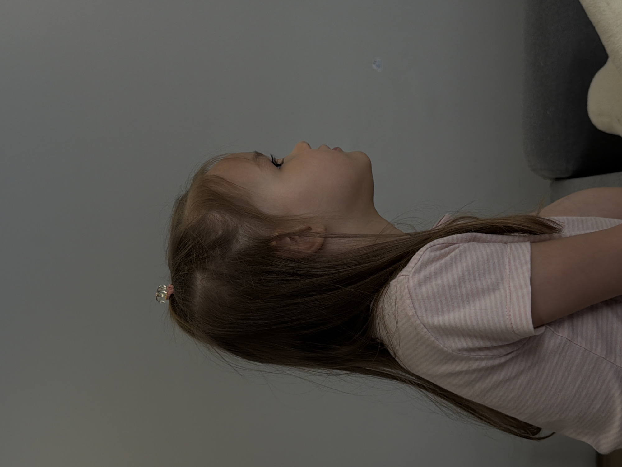 A young child is pictured in profile, looking upward. She has fair skin and light brown hair pulled partially back with a decorative hair clip. She is wearing a pink and white striped t-shirt. The background is a gray wall. A portion of a gray couch with a white stuffed animal is visible in the upper right corner. The image's location is not discernible from the provided information.