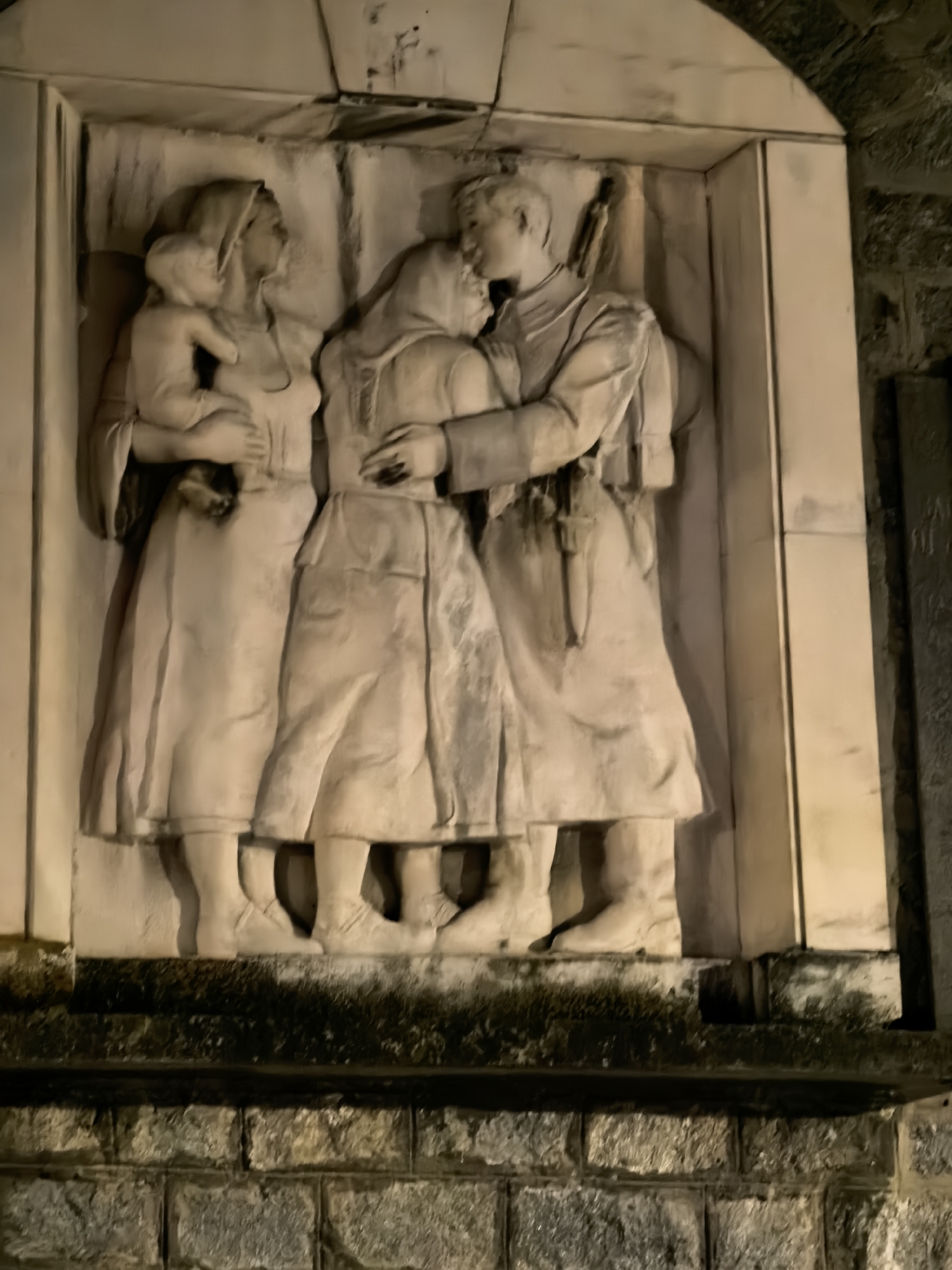 This image depicts a bas-relief sculpture, potentially a war memorial, set into a stone wall. The sculpture portrays three figures: a woman holding a child, another woman, and a soldier. The soldier, positioned on the right, is embracing the other woman. He appears to be wearing a military uniform with a rifle slung over his shoulder. The woman on the left is holding a baby and looks toward the embracing couple. The relief is framed by a rectangular structure. The wall beneath the sculpture is constructed of stone blocks. The image location is listed as Unknown, Unknown.