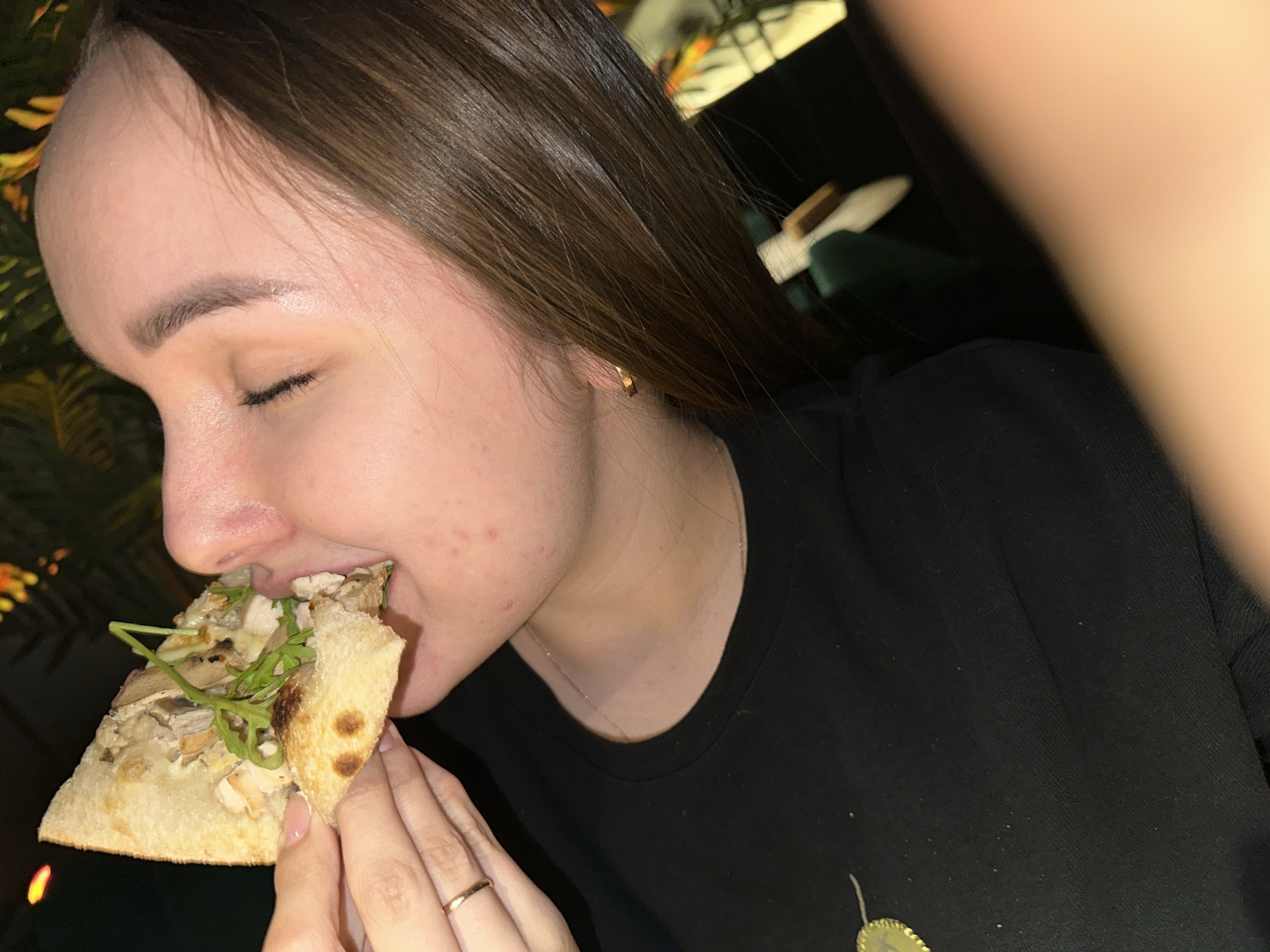 A woman is eating a slice of pizza. She has brown hair, fair skin, and a gold hoop earring. She is wearing a black shirt. The pizza slice appears to contain mushrooms and some green leaves on top. The scene is dimly lit. The location is not clear from the image, and there is no information to suggest the city of Chaikovsky, Russia.