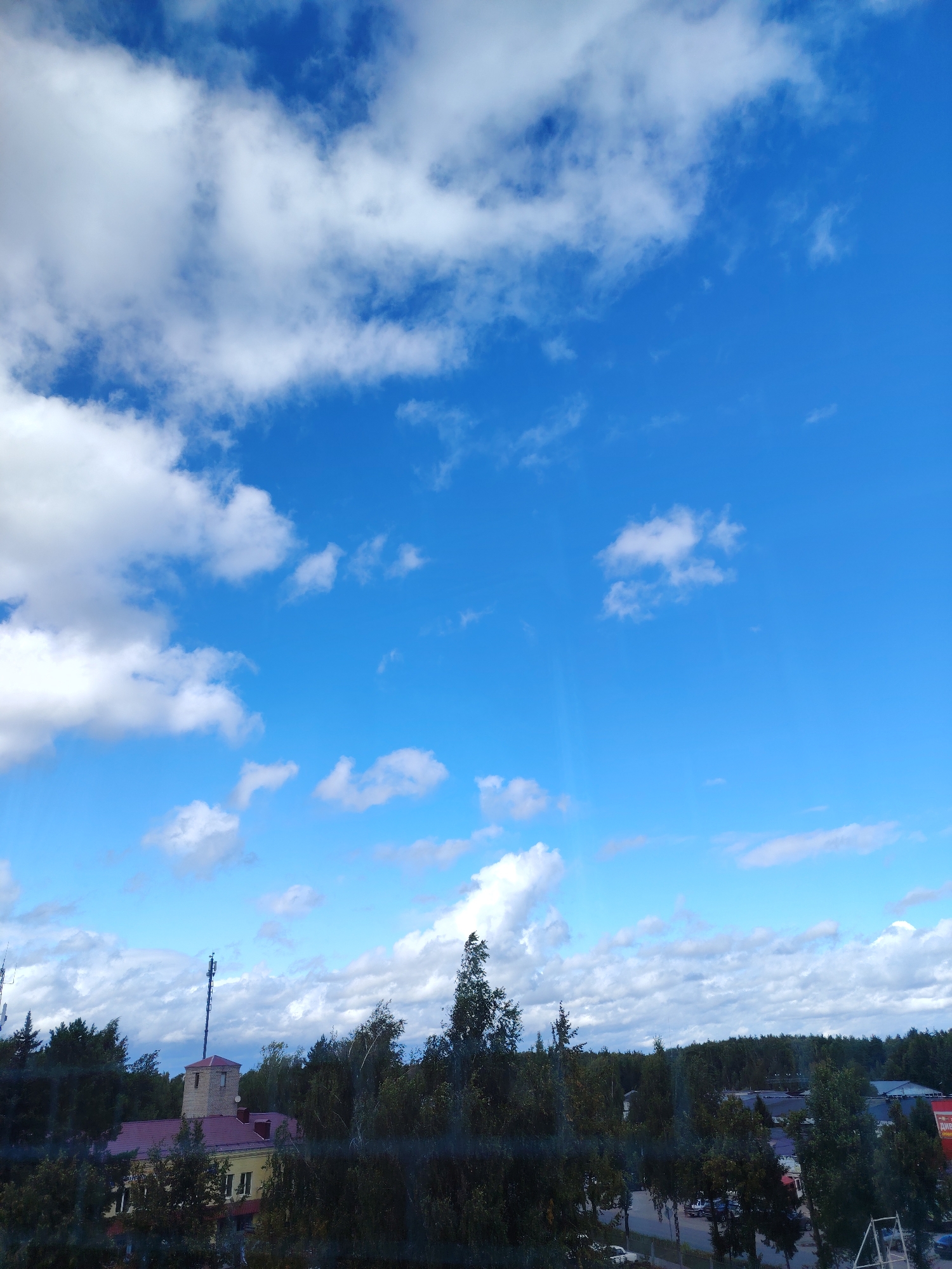A bright blue sky with scattered white cumulus clouds dominates the upper portion of the image. Below the sky, a dense line of dark green trees forms a horizon. In the mid-ground, a building with a red roof and yellow facade is visible, topped by a square brick tower with a tall antenna. A cell tower stands prominently behind the tower. In the foreground, a dark, blurred barrier, possibly a window with a screen or a fence, obscures a view of a street with parked cars and other buildings. The scene appears to be an outdoor urban environment in Konakovo, Russia, during the daytime.