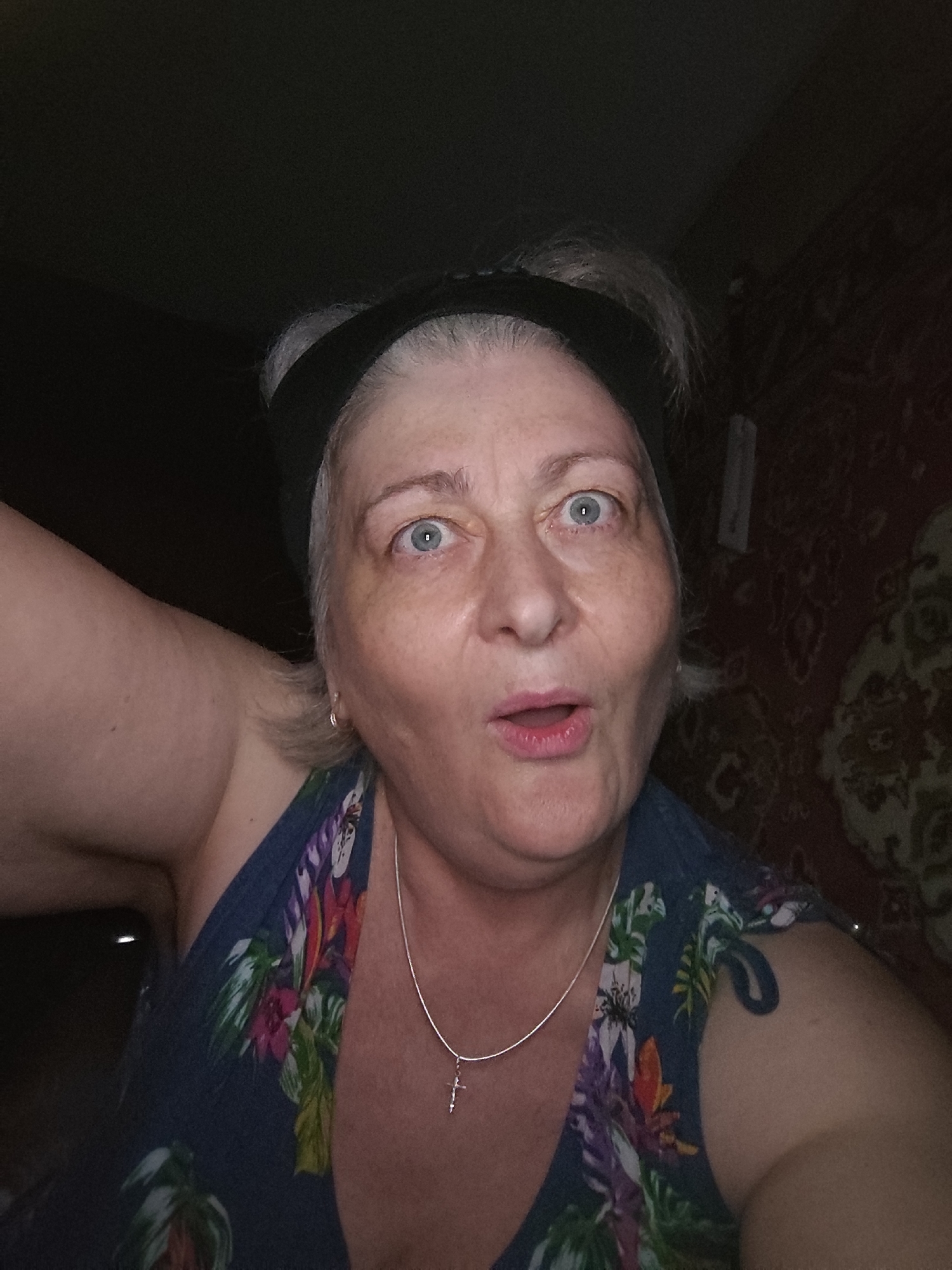 The image displays a close-up, dimly lit indoor selfie of an adult female. Her grey hair is pulled back with a black headband, revealing wide blue eyes and a slightly agape mouth, indicative of a surprised or expressive facial gesture. She wears a dark blue sleeveless top featuring a vibrant floral pattern. A thin silver-colored necklace with a small cross pendant hangs around her neck, and a simple earring is visible in her left ear. Her left arm and shoulder are prominent on the left side of the frame. The background on the left is dark and indistinct, while the right side reveals a dark red patterned wall with a repeating motif and a faint vertical light-colored object in the upper section. The setting is an interior space.