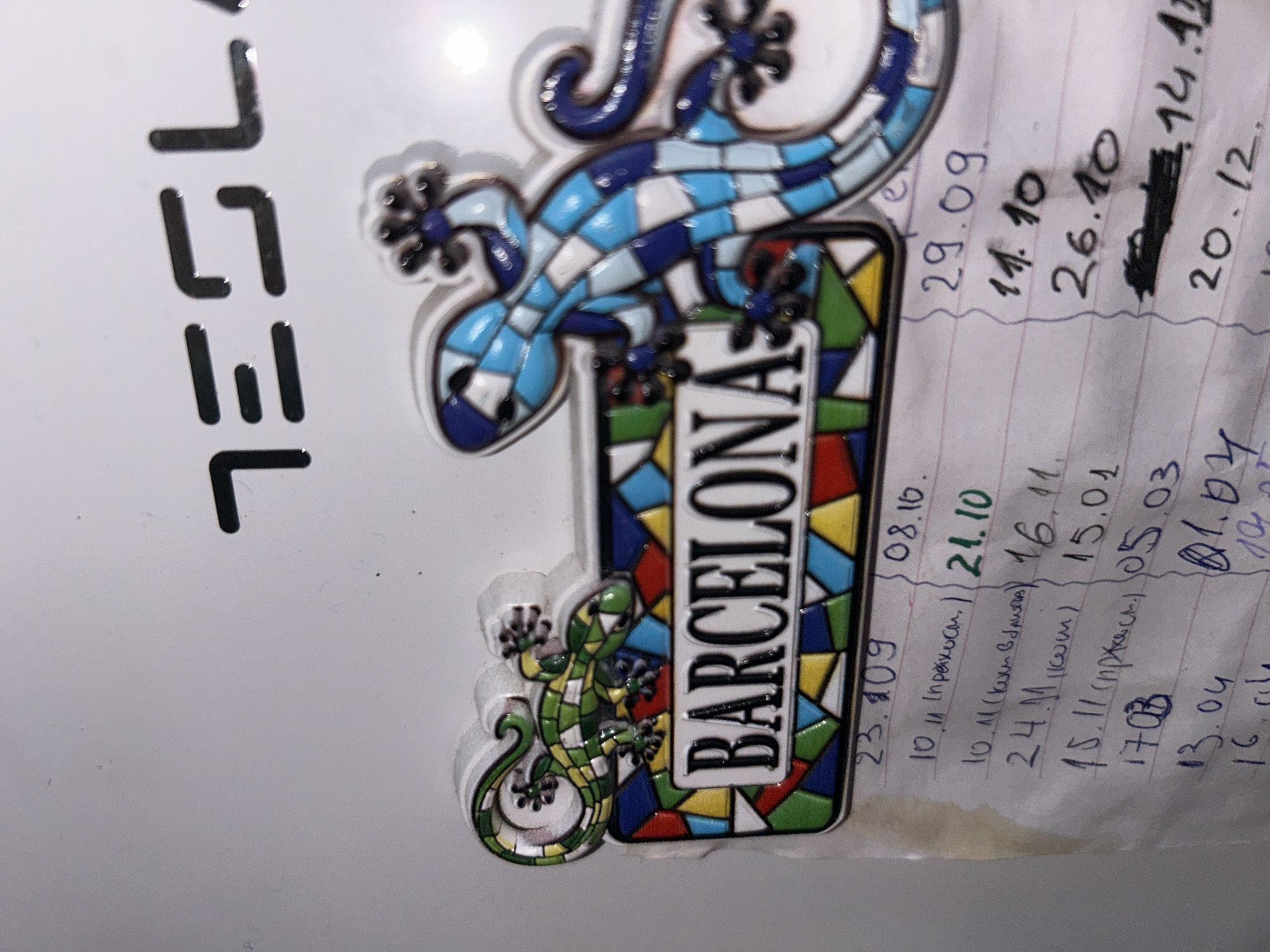 The image shows a section of a white refrigerator door, likely in an apartment or household setting, featuring a Tesla logo and two decorative magnets. One magnet is a colorful gecko on a stained-glass-like background with the word "BARCELONA" written on it. The other magnet is a larger, blue and white mosaic gecko. Below the magnets, a handwritten list on lined paper is visible, containing dates and what appear to be personal notes or entries. The Tesla logo is a metallic, stylized text. The lighting suggests an indoor environment, with some glare on the refrigerator door. There are no people present in the image, and no identifiable landmarks or context that would suggest a specific location in Sofia, Bulgaria. The image is a close-up, focusing on the magnets and the written list.