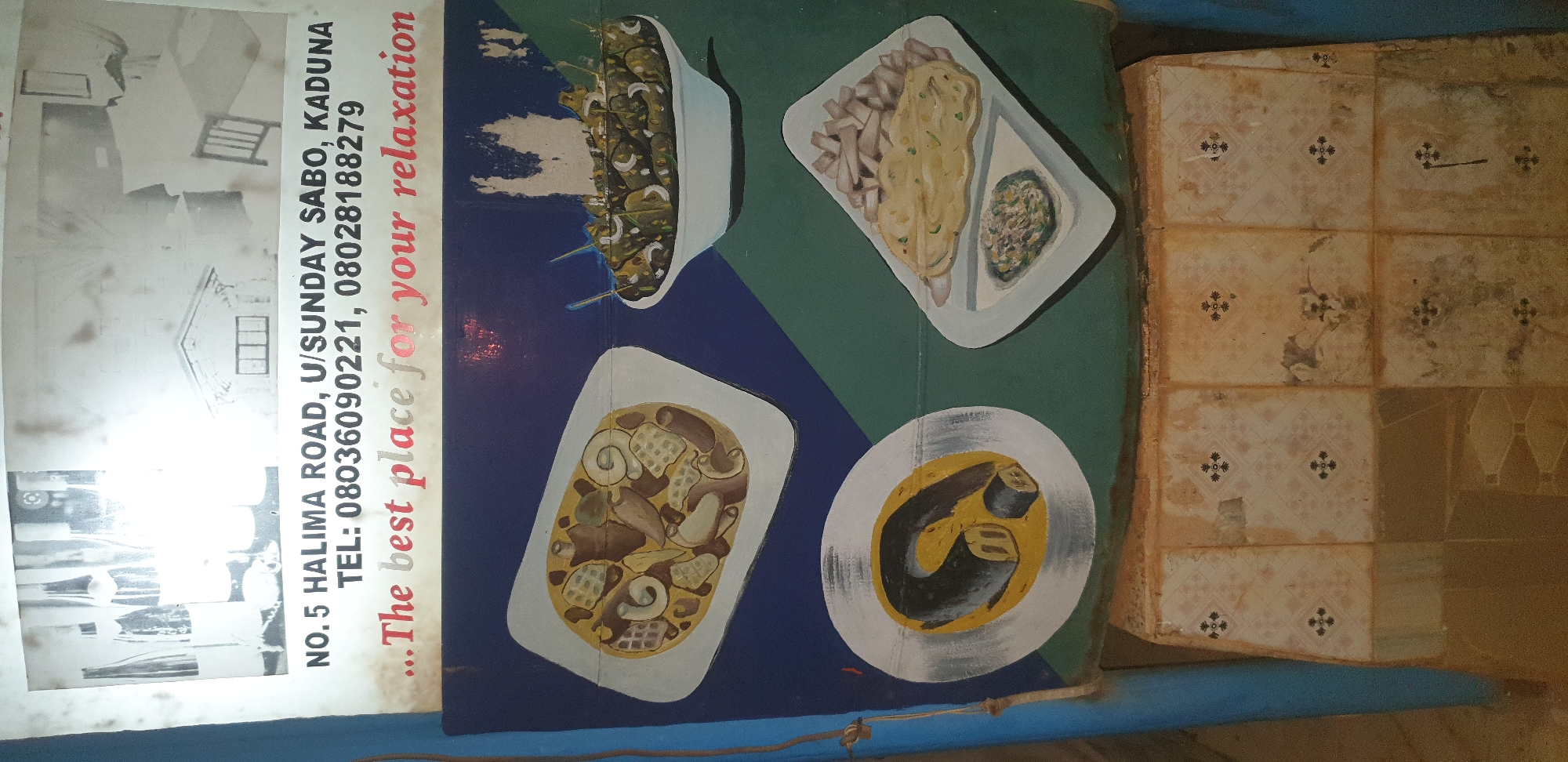 This image shows a faded, hand-painted advertisement for a business located at NO. 5 HALIMA ROAD, U/SUNDAY SABO, KADUNA, NIGERIA. The advertisement features illustrations of food items and text promoting the business as "...The best place for your relaxation." The phone numbers listed are 08036090221 and 08028188279. The advertisement is mounted on a wall above a tiled surface. The overall scene suggests an outdoor or semi-outdoor setting, possibly a shop or restaurant entrance. The art style is naive and illustrative.