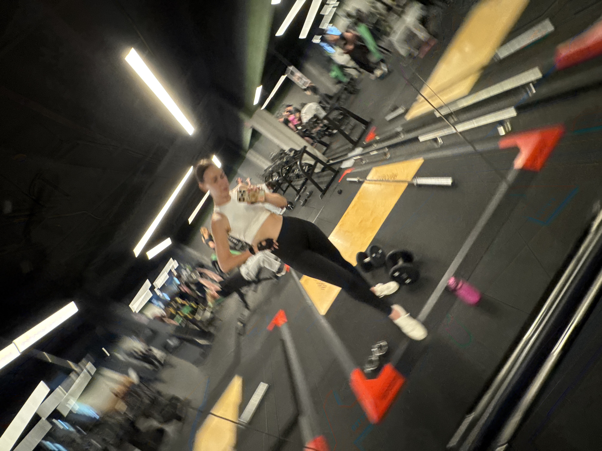 The media file depicts a young woman taking a selfie in a gym. She is wearing a white crop top and black leggings, with white sneakers. The gym appears to be well-equipped with various weight machines and free weights, including dumbbells. The lighting is bright, with fluorescent lights visible on the ceiling. The image is taken from a high-angle perspective, capturing the woman and a portion of the gym floor. The background shows other gym-goers, some engaged in workouts. There are also red markers on the floor, possibly indicating training zones or equipment placement. The overall atmosphere seems energetic, consistent with a fitness environment. The motion blur suggests the photo was taken quickly or with movement. The location is identified as Sochi, Russia. No specific time of day or weather conditions are discernible. There is no visible text in the image.
