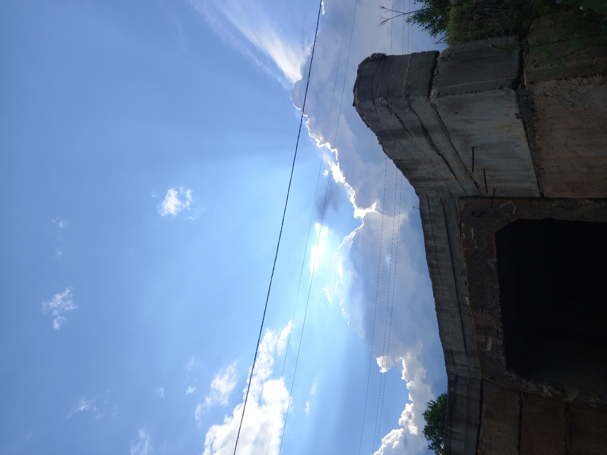 This low-angle, landscape-oriented photograph shows a section of a concrete structure against a bright blue sky. The concrete structure appears to be part of an overpass or bridge, with multiple layered sections visible. The rough texture and visible rebar suggest it is a utilitarian construction. In the sky, scattered white clouds are present, with some large cumulus clouds catching the sunlight. Rays of light are fanning out from behind one of these clouds, indicating a bright, sunny day. Several power lines are strung across the frame, running diagonally and intersecting with the concrete structure and the sky. Vegetation, appearing as green foliage and dry branches, is visible at the top edge of the image, peeking from behind the concrete. The overall scene evokes a sense of an outdoor, possibly rural or semi-urban environment. The time of day is likely midday given the brightness of the sun and the sharp contrast. There is no visible text in the image.