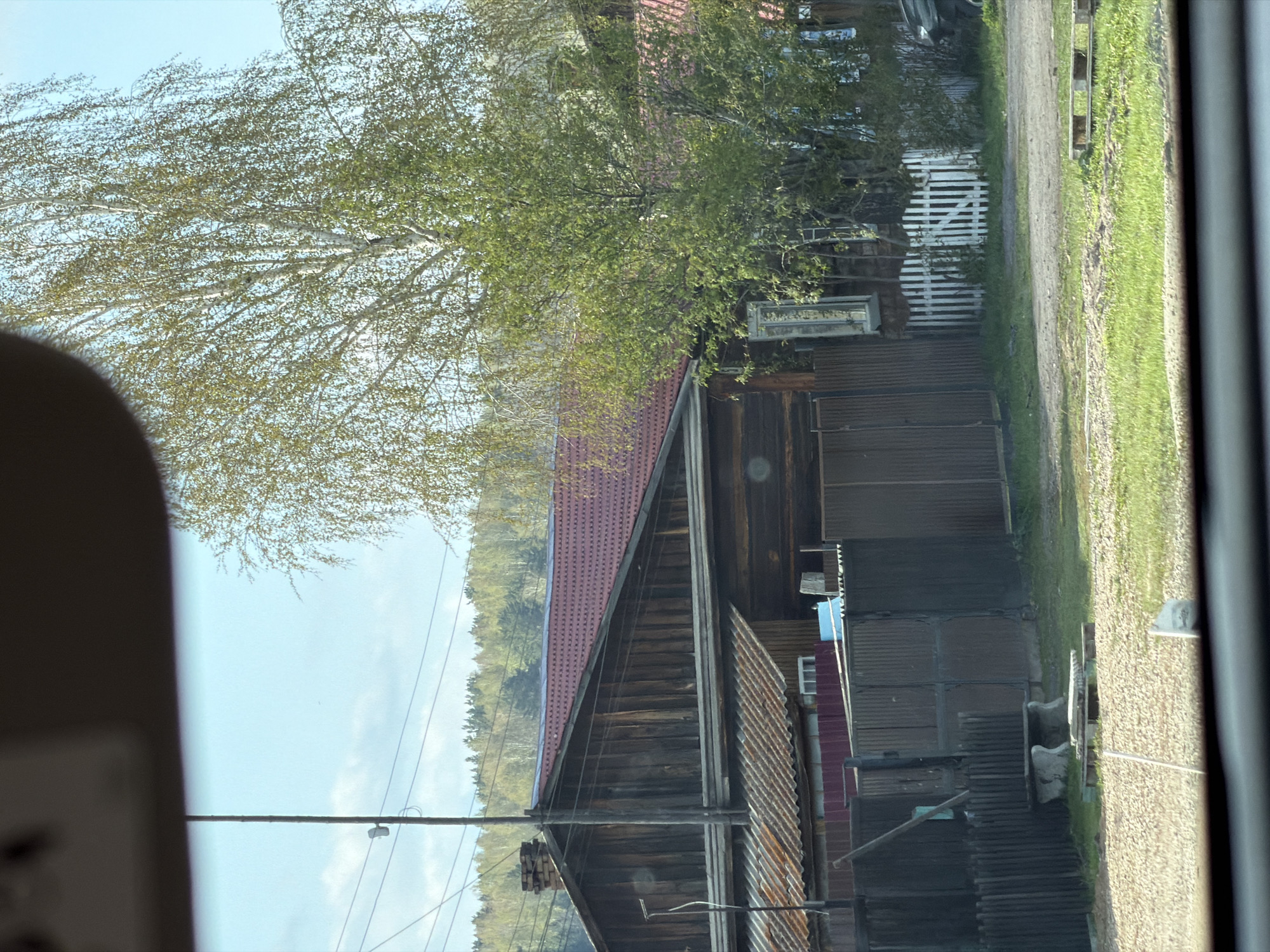 The image shows a view from inside a vehicle or building looking out onto a rural or suburban scene. There is a wooden building with a red roof visible in the center of the frame. The building appears to be old and possibly weathered. A large tree with thin branches and green leaves obscures part of the building and extends towards the top of the image. A road or path runs alongside the building, with grass and vegetation lining the edge. A white fence can be seen near the building. Power lines are strung across the sky. The sky is blue with light clouds. The location appears to be outside a city, given the type of architecture and landscaping.
