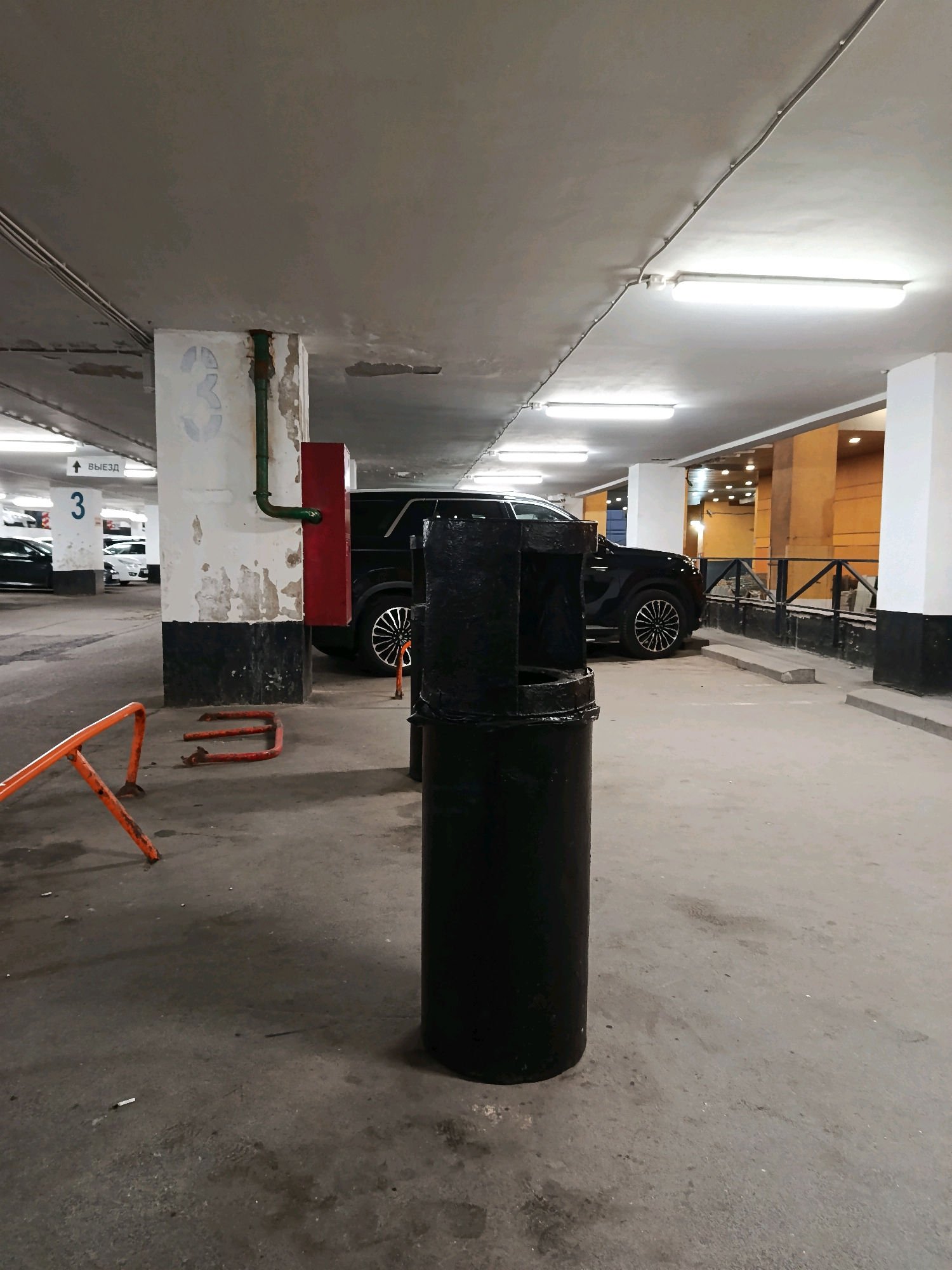 An interior shot of a dimly lit, underground parking garage shows a black trash can in the foreground. The trash can is cylindrical and appears to be made of metal. Behind the trash can, a black SUV is parked. The garage features concrete pillars, fluorescent lighting, and a painted number "3" on one of the pillars. An orange barrier is visible to the left of the trash can, and a red fire extinguisher box is mounted on a pillar behind the trash can. The floor is concrete and shows signs of wear and tear, with some water stains and what appear to be cigarette butts. The overall atmosphere is utilitarian and industrial.