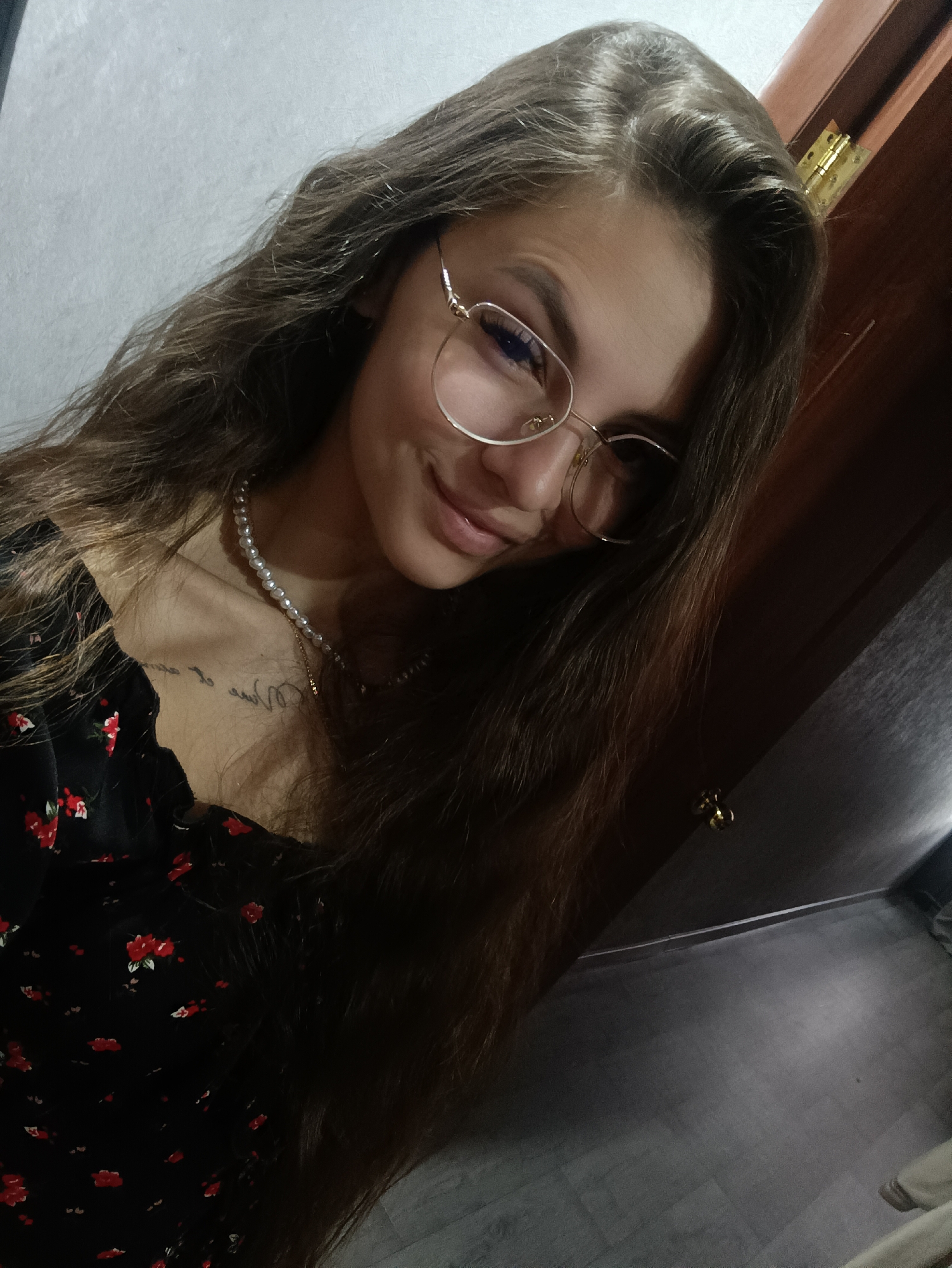 A medium close-up shot shows a young woman with long, wavy brown hair, wearing gold-rimmed glasses, a pearl necklace, and a black dress with small red floral patterns. A glimpse of a tattoo is visible on her collarbone. Her head is tilted slightly to the side with a small smile. The background includes a white wall and a dark wooden door with a gold-colored handle. The floor appears to be a dark wood or laminate. The location is not discernible.