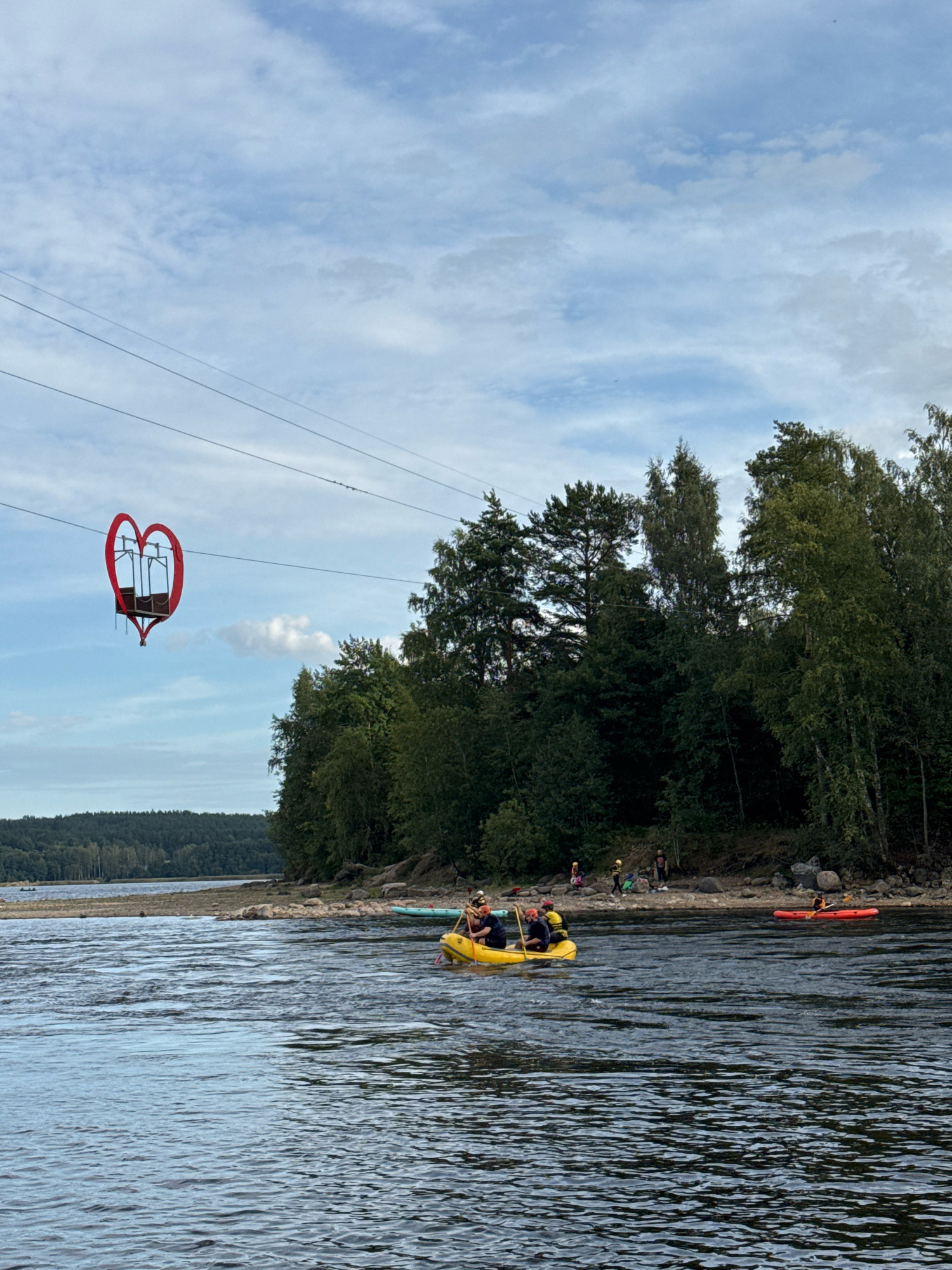 This image depicts a scenic view of the Don River in Losevo, Russia, on a cloudy day. In the foreground, the wide expanse of the Don River shows moderate ripples and waves, reflecting the overcast sky. On the water, a yellow raft is being paddled by several individuals wearing life vests and helmets. Further to the right, a person is kayaking in a red kayak.
Along the riverbank, which is rocky and covered with sparse vegetation, a group of people are gathered. Behind them, a dense line of green trees forms the backdrop, with taller pines and deciduous trees visible.
Suspended in the sky by two taut wires, a unique attraction is visible: a red, heart-shaped gondola. This gondola, likely part of a cable car system, hangs suspended above the river, adding a distinctive element to the landscape. In the distance, a forested hillside slopes down towards the river, with a calm body of water visible on the far left. The overall atmosphere is one of outdoor recreation and natural beauty.