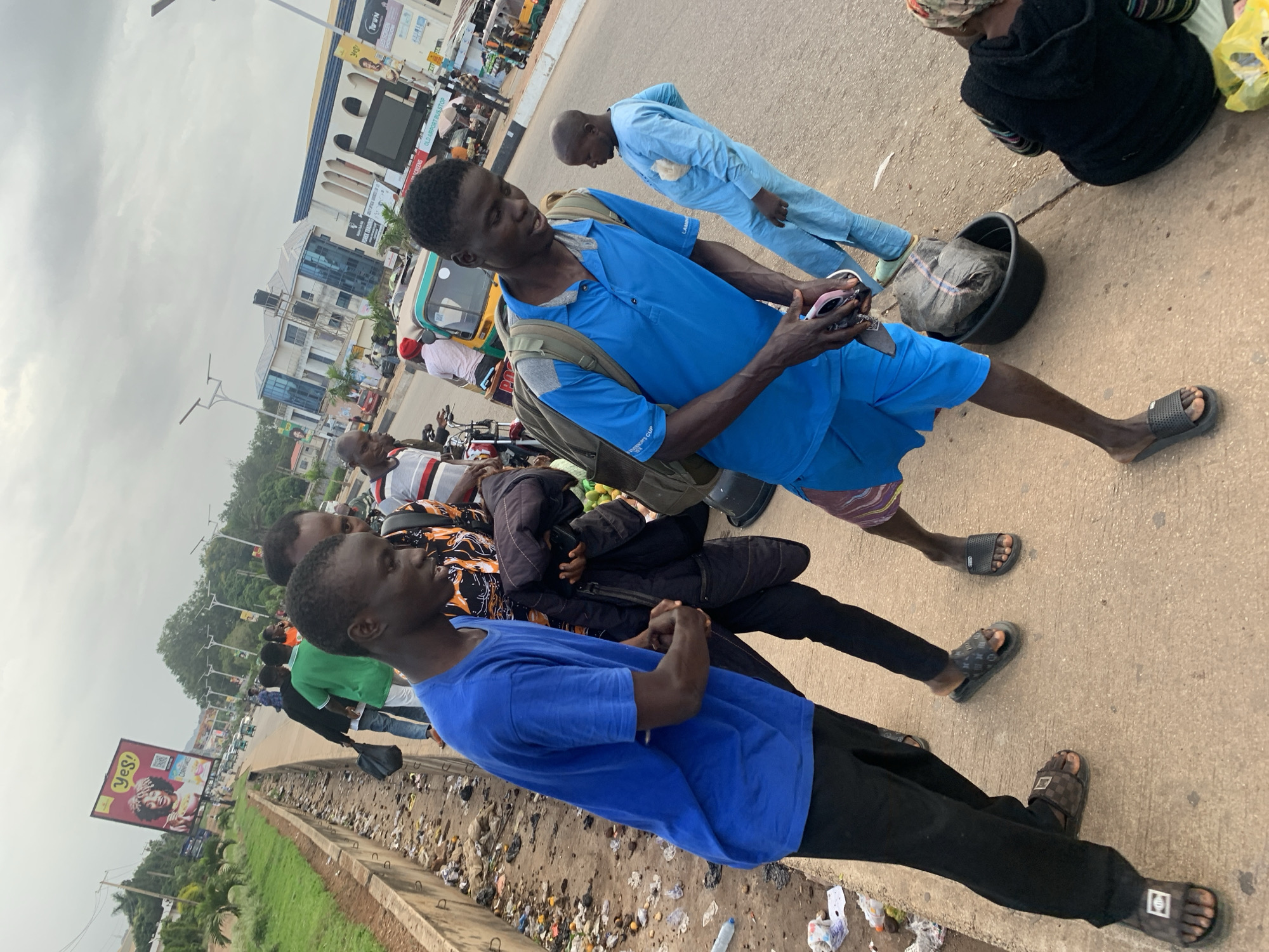 In Barakin Lerawi, Nigeria, a group of young men are gathered on a sidewalk next to a busy road. The sidewalk is lined with a concrete barrier and strewn with litter. In the foreground, two young men are walking. One, wearing a blue t-shirt and black pants, carries a dark jacket over his arm. The other, in a blue polo shirt and blue shorts, has a backpack slung over his shoulder and is holding a phone. Further back, another young man in a light blue traditional garment stands on the road, facing away from the camera. In the background, a yellow auto-rickshaw, motorbikes, and other vehicles are visible on the road. Buildings and billboards, including one advertising "YES!" cereal, line the opposite side of the street. The sky is overcast.
